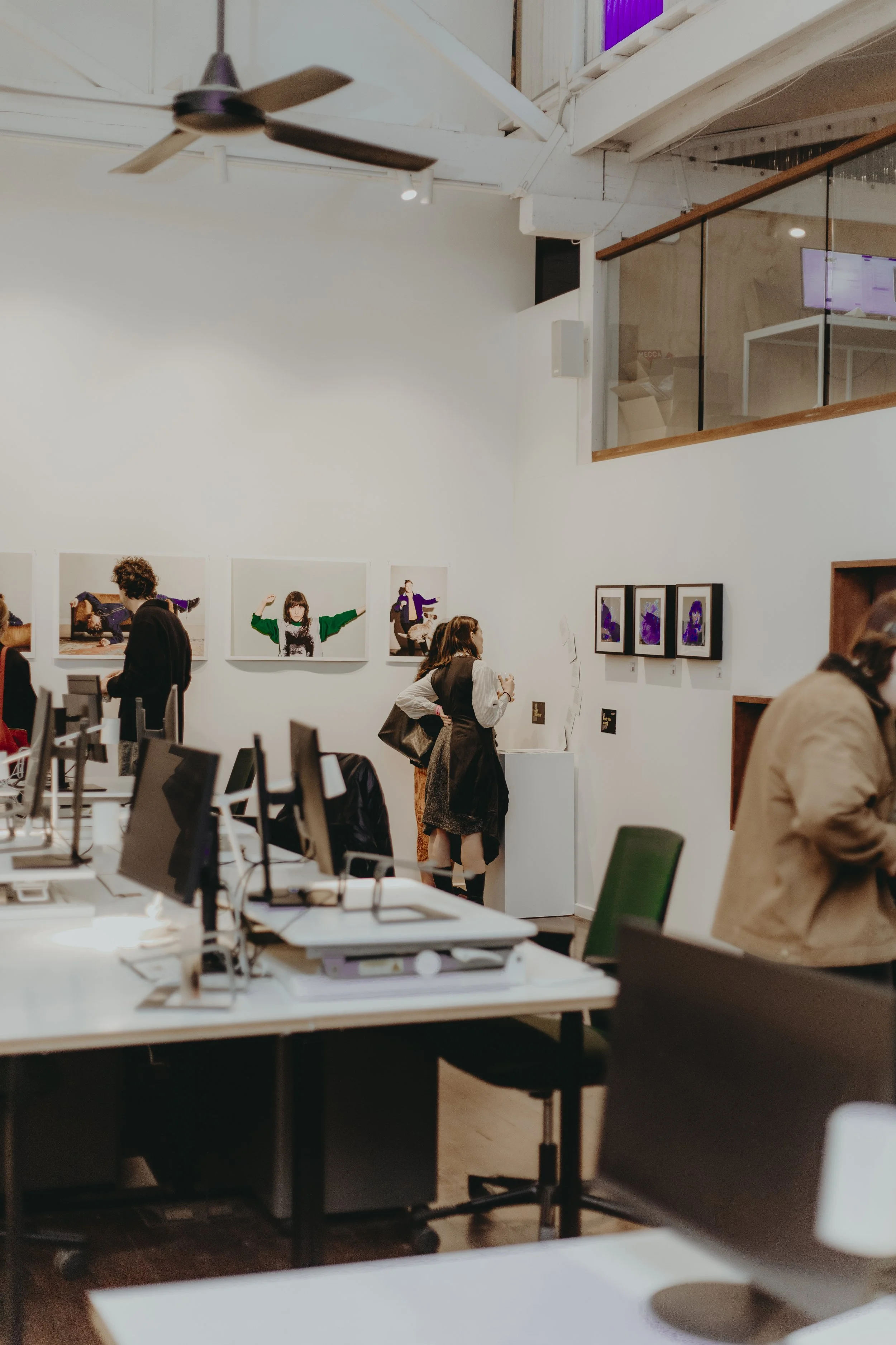 An office or gallery space with people viewing artwork on the walls, multiple computer desks with monitors, and a high ceiling with exposed beams and a ceiling fan.