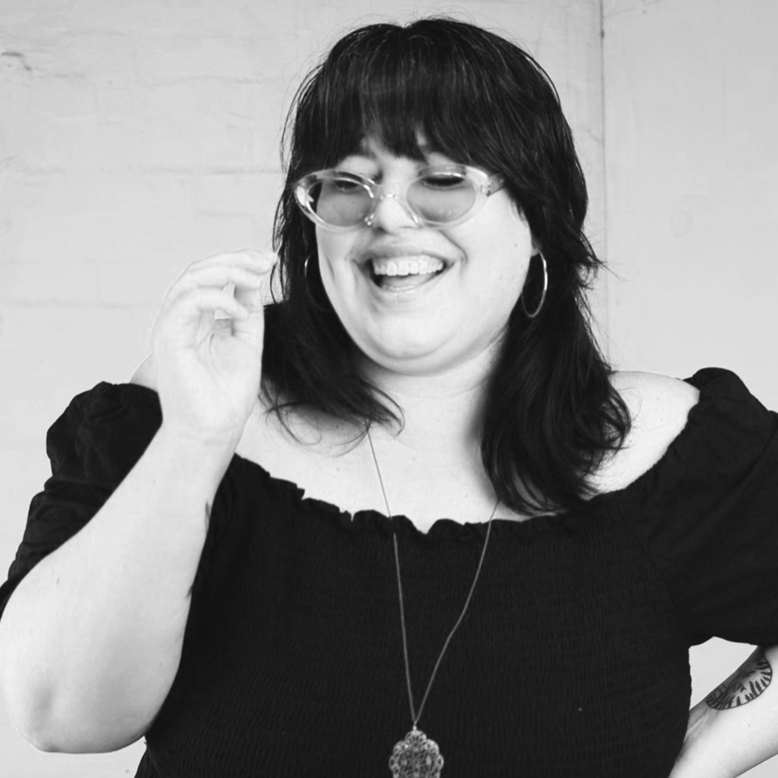 A woman with dark hair, wearing glasses and hoop earrings, smiling and adjusting her glasses, against a plain wall background, wearing a black off-the-shoulder top and a long necklace.