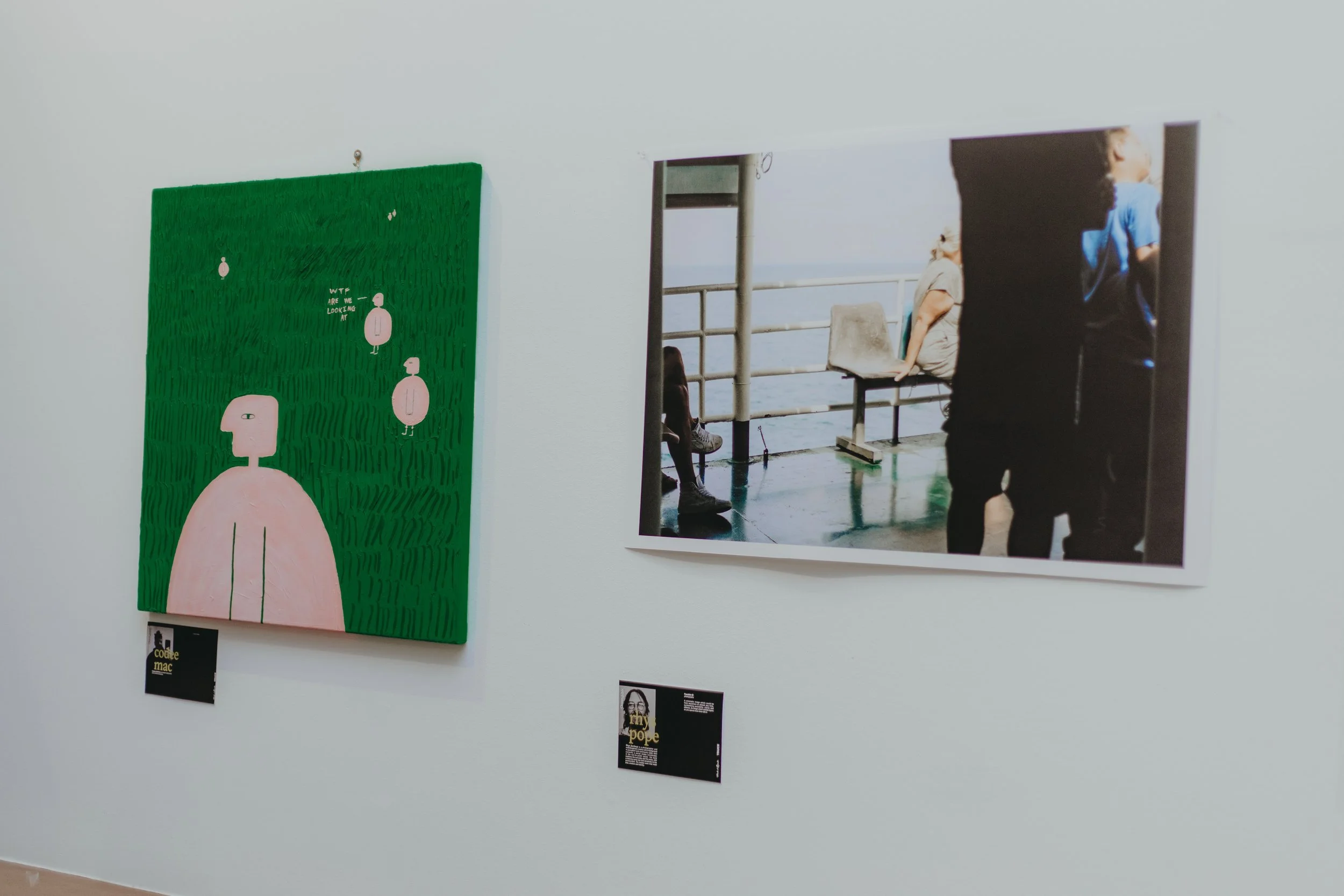 An art gallery wall featuring a green painting with abstract figures and a black label, next to a photograph of people sitting on a railing on a building with a view of the ocean.