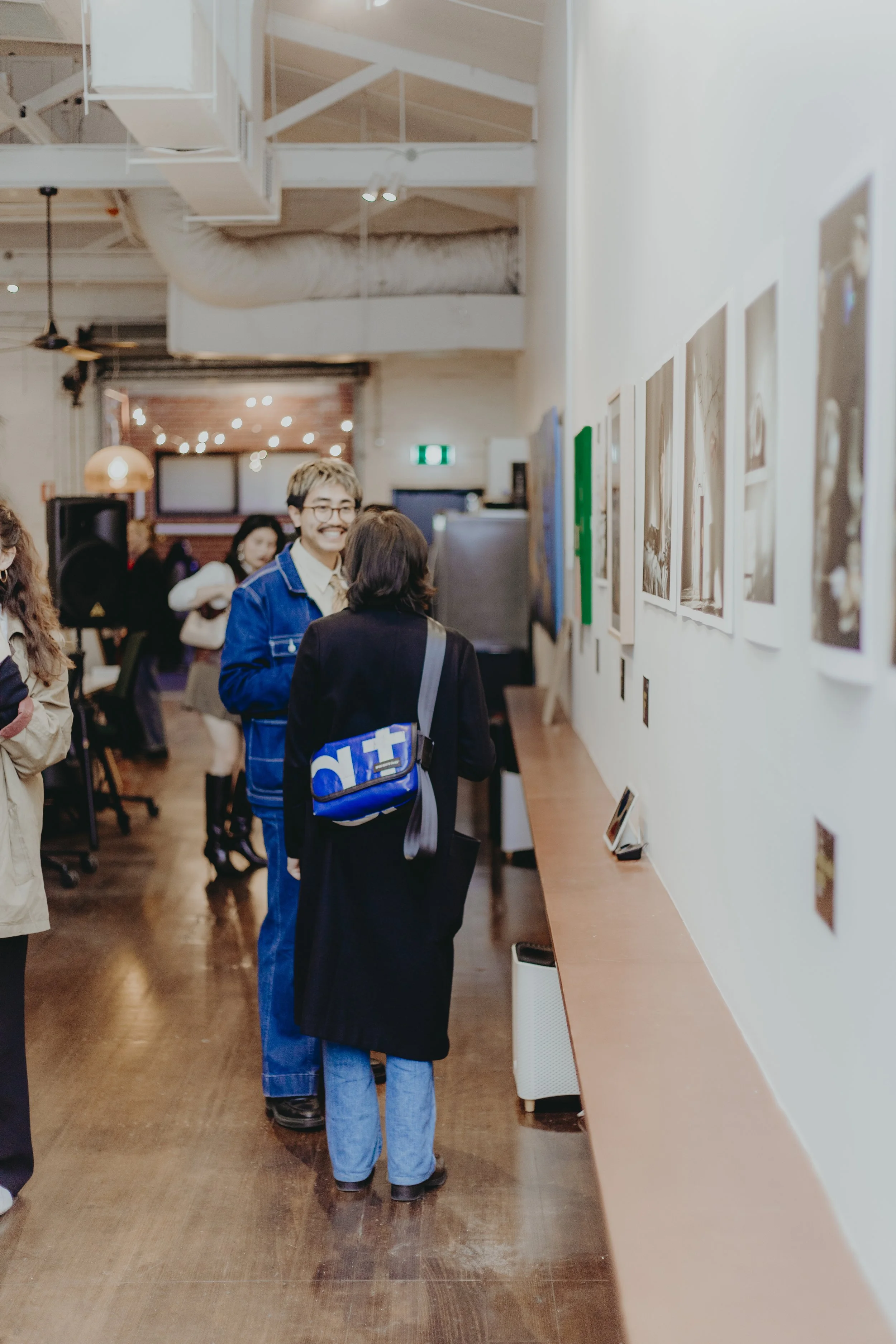 Gallery with group of people viewing artworks displayed on white walls, wooden floors, and soft lighting.