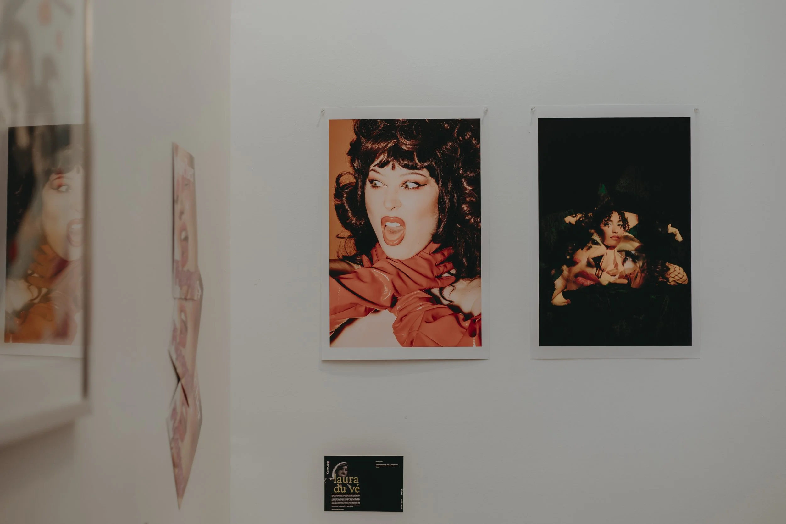 Photograph of an art exhibition on a white wall featuring portraits of women. The central image shows a woman with voluminous curly hair and a surprised expression, wearing a red outfit. To the right is another woman with curly hair, wearing a hat. T