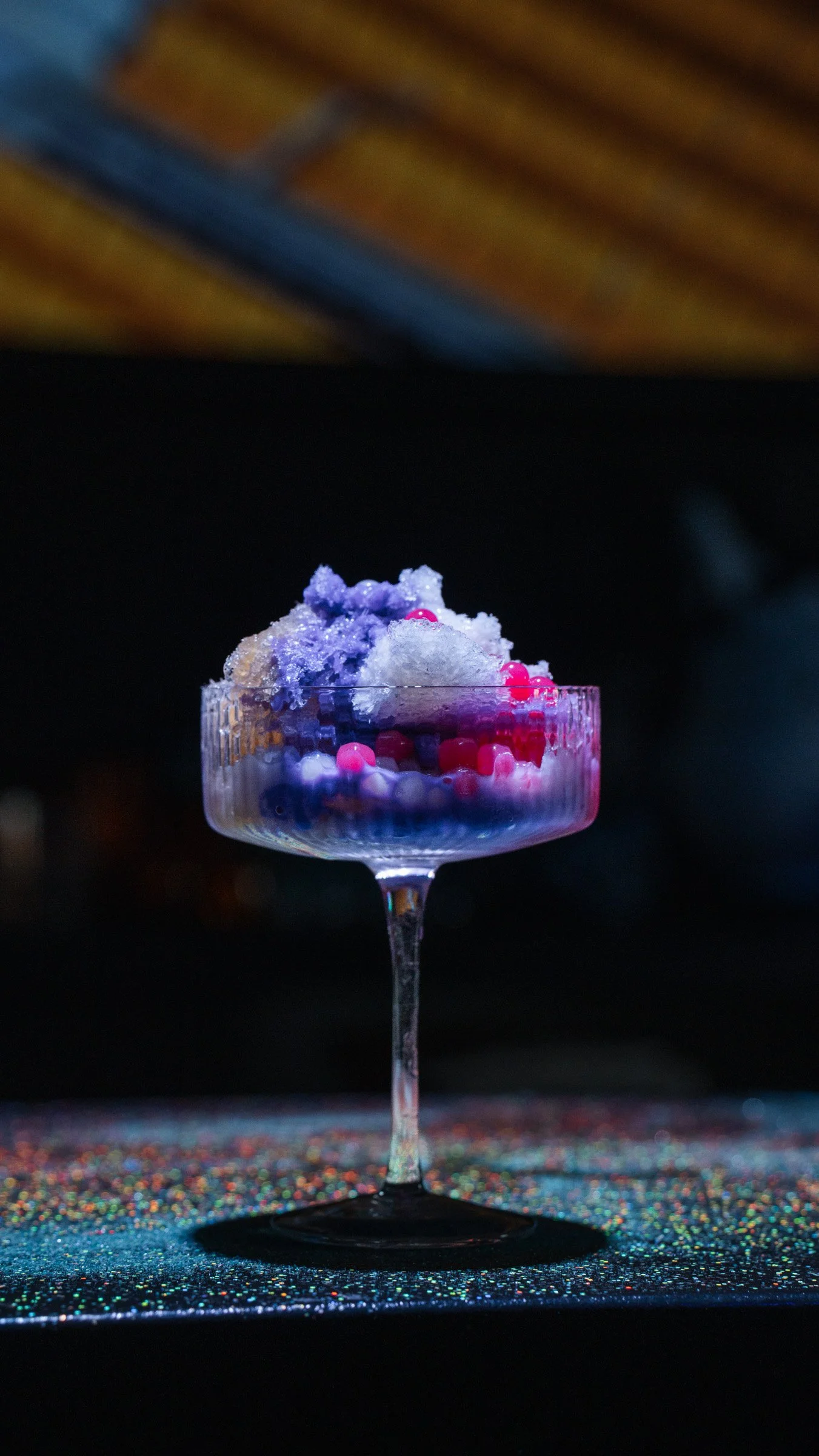 A cocktail glass with a colorful, layered dessert topped with cotton candy and small pink and purple spheres, illuminated by vibrant lighting.