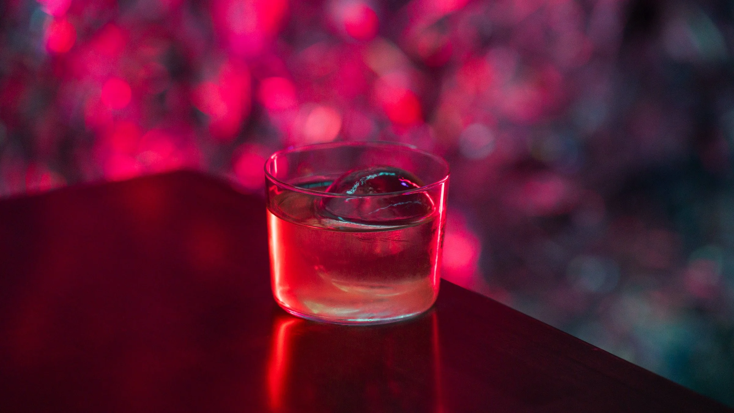 A glass of water with ice cube on a dark table against blurred pink and purple bokeh background.