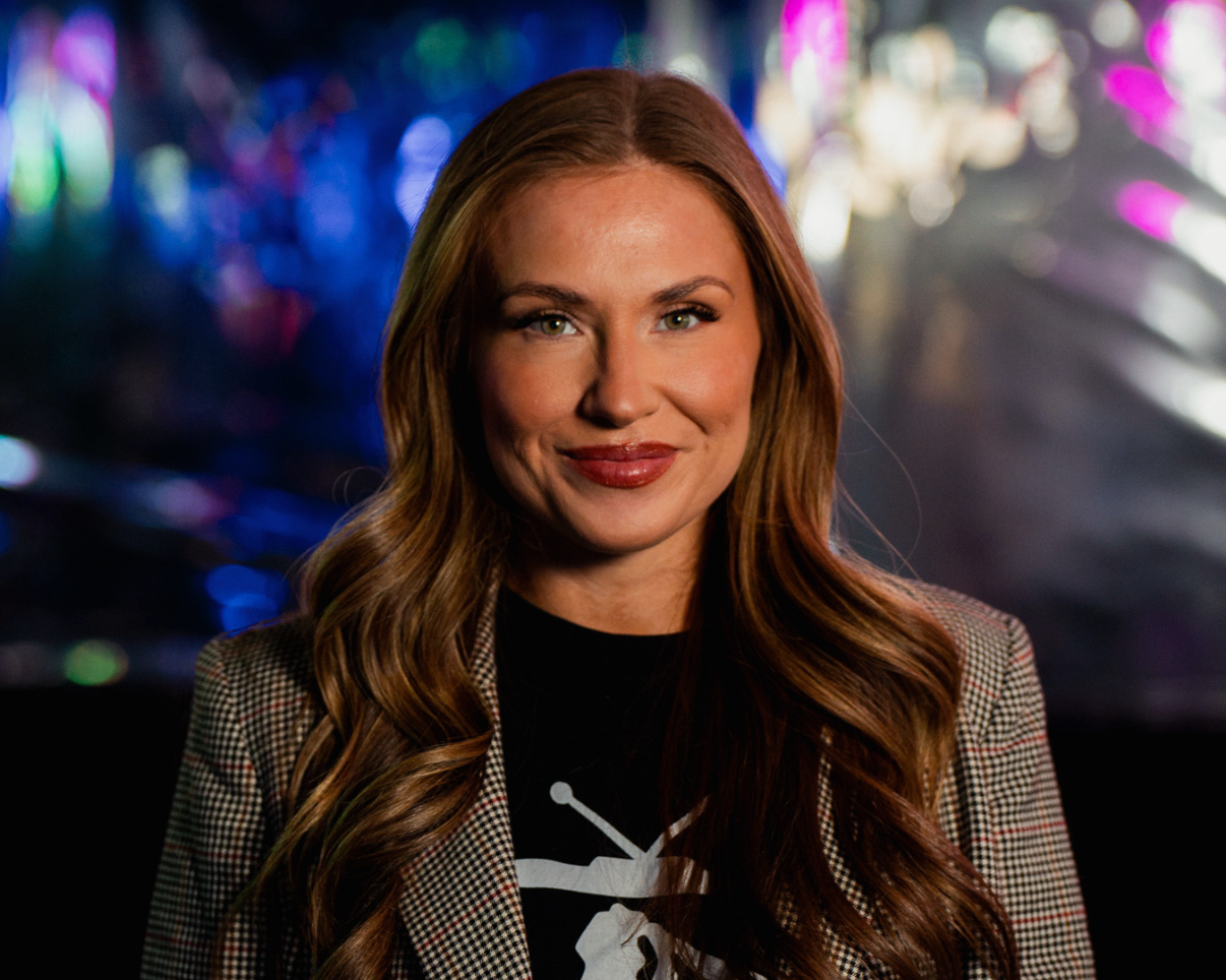 A woman with long wavy auburn hair and fair skin smiling at the camera, wearing a checked blazer over a black t-shirt with a white skull graphic, against a colorful blurred city lights background.
