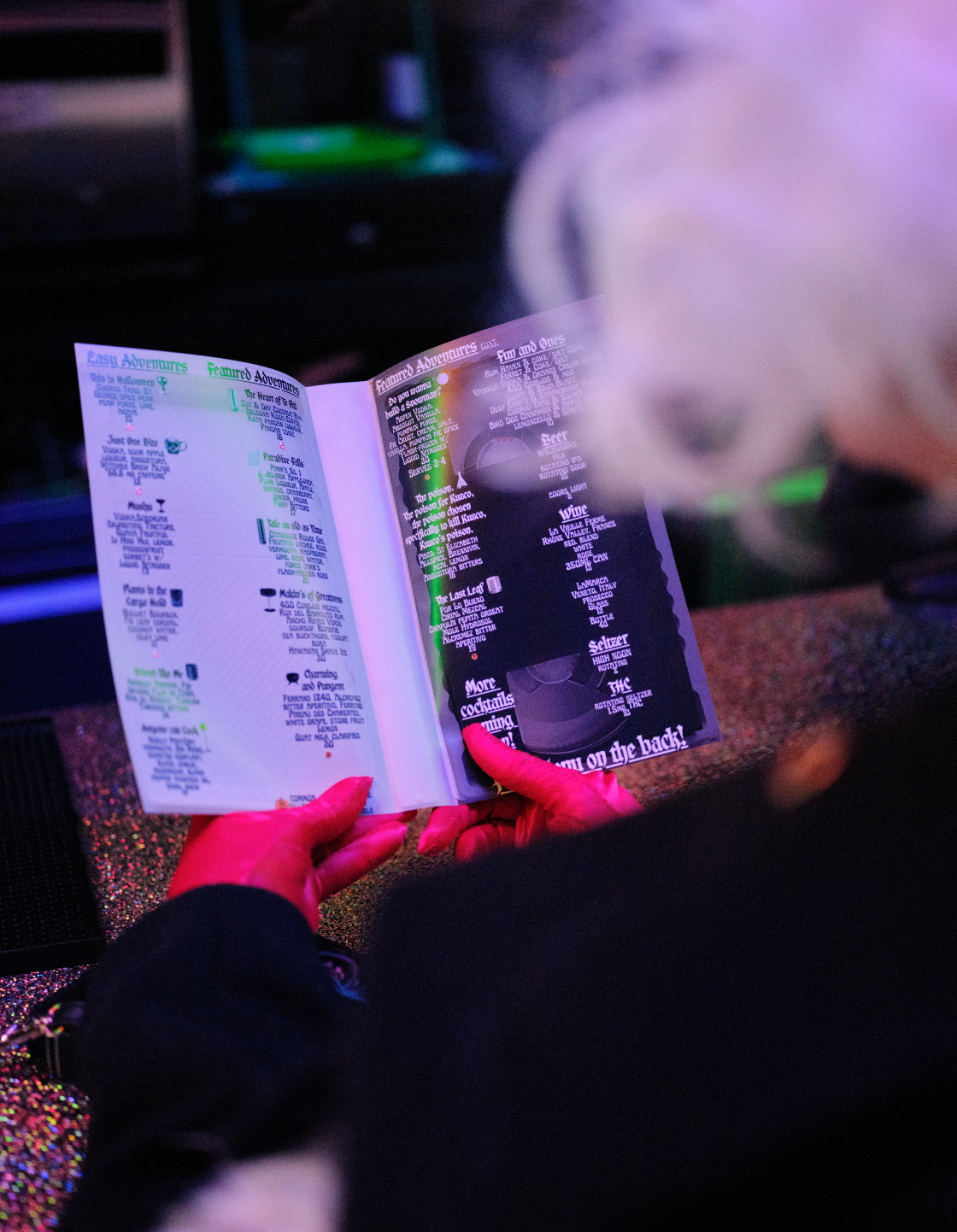 Person holding a black-lit menu with neon signs, reading a drink menu in a dimly lit venue with colorful lighting.