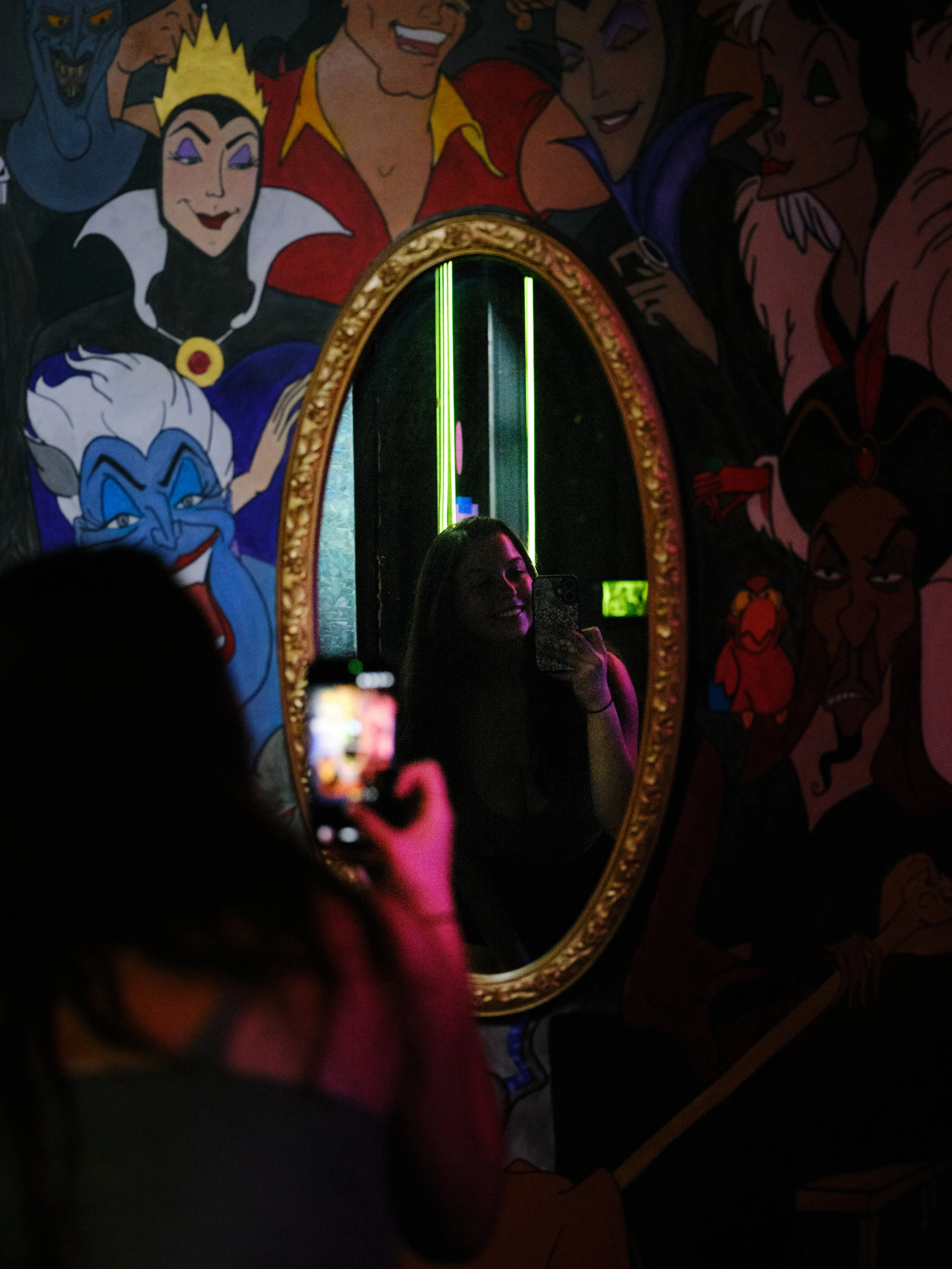 A woman taking a selfie in front of an ornate mirror with a Disney villain mural background, featuring characters like Ursula and others, with colorful lighting.