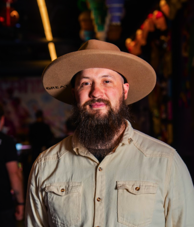 A bearded man wearing a wide-brimmed hat and a beige button-up shirt standing in an indoor setting with colorful lights and decorations in the background.