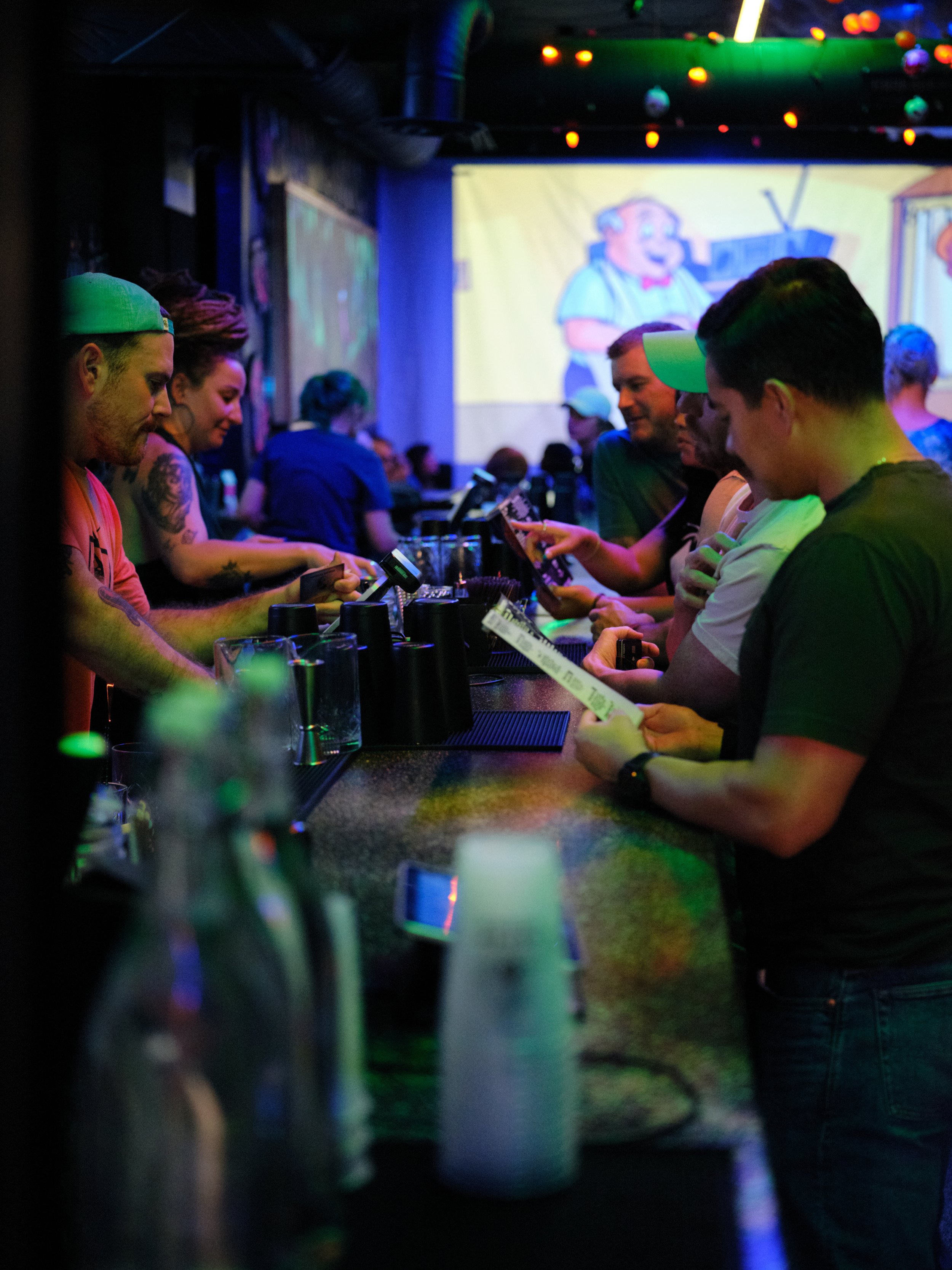 People gathered around a bar counter in a dimly lit room with colorful lighting, with a large animated cartoon character projected on the wall in the background.