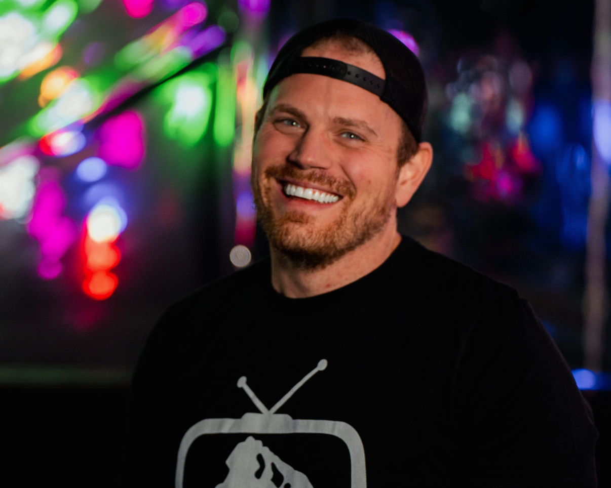 A smiling man wearing a black cap backwards and a black t-shirt with a white logo of a mountain and cell phone towers, standing in front of colorful blurred neon lights.