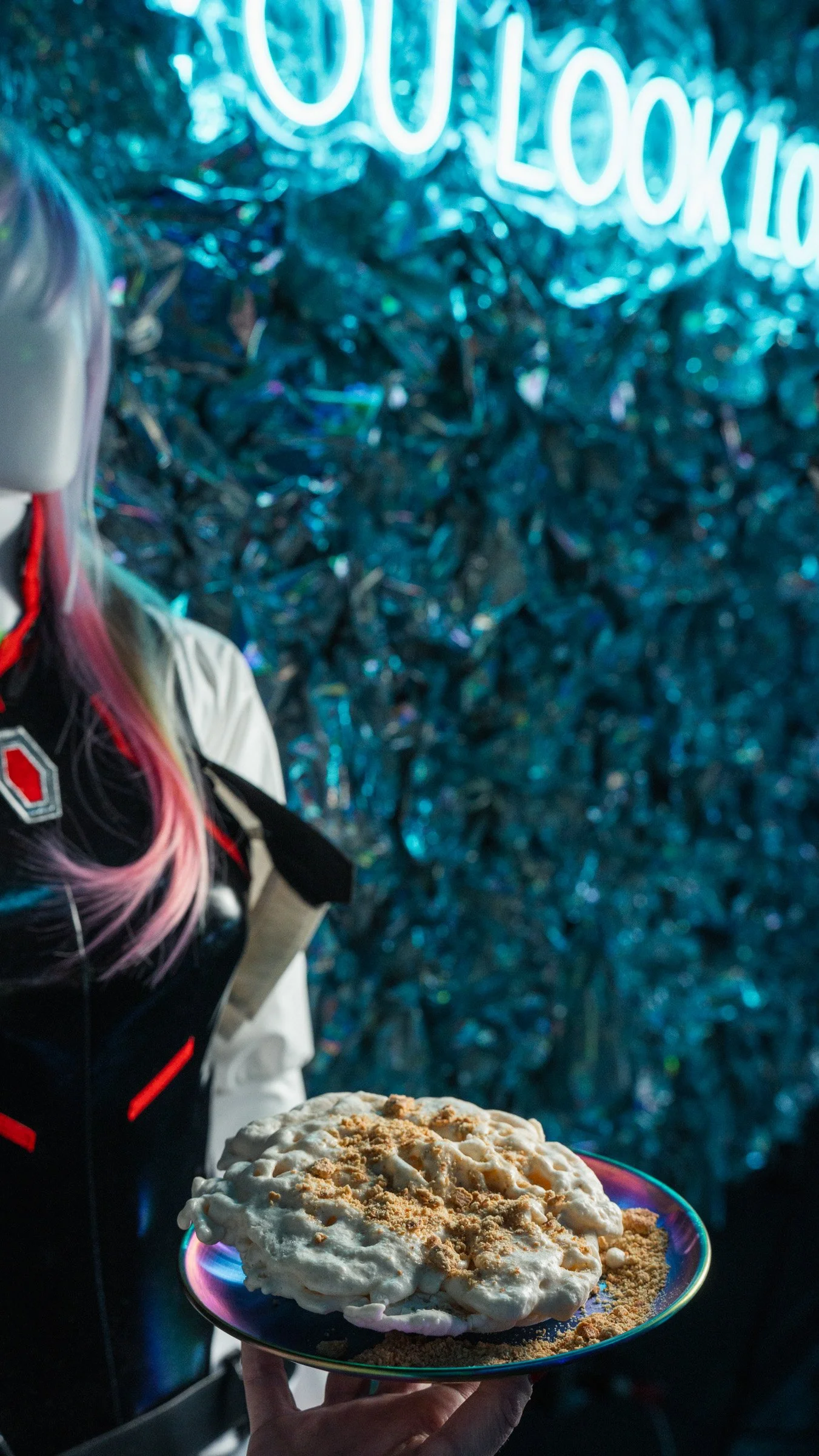 A person holding a plate with a large serving of ice cream topped with crumbs, in front of a metallic, shiny background and a blue neon sign.