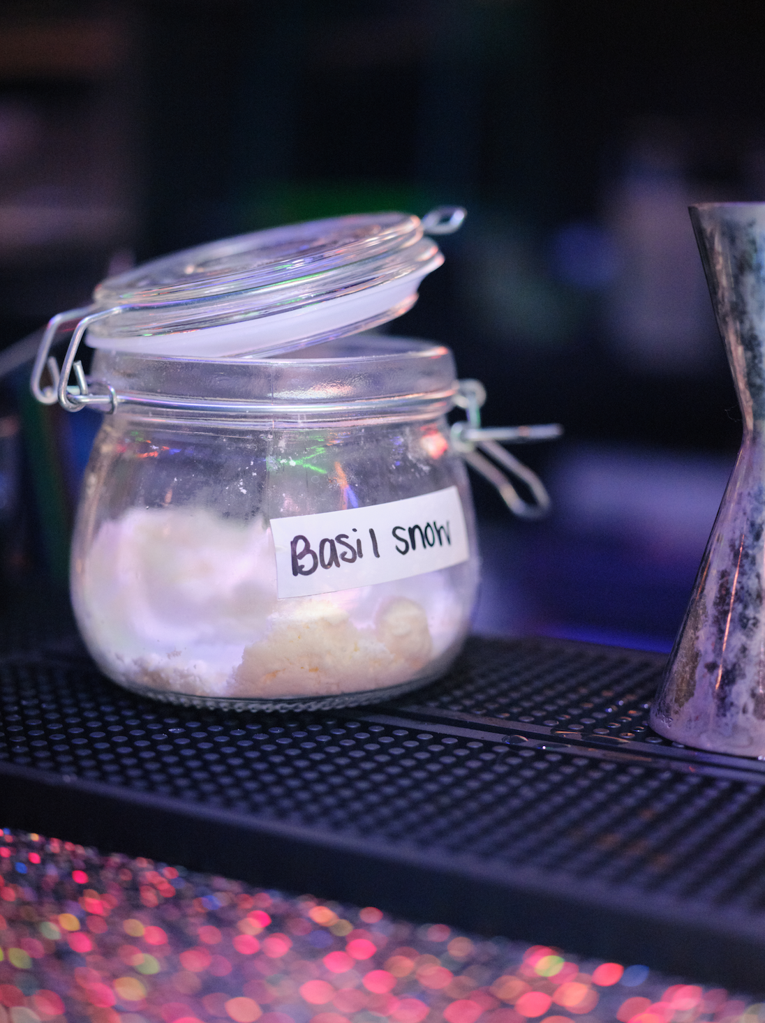 A glass jar labeled 'Basil Snow' containing a white powdered substance, placed on a black textured surface with colorful lights in the background.