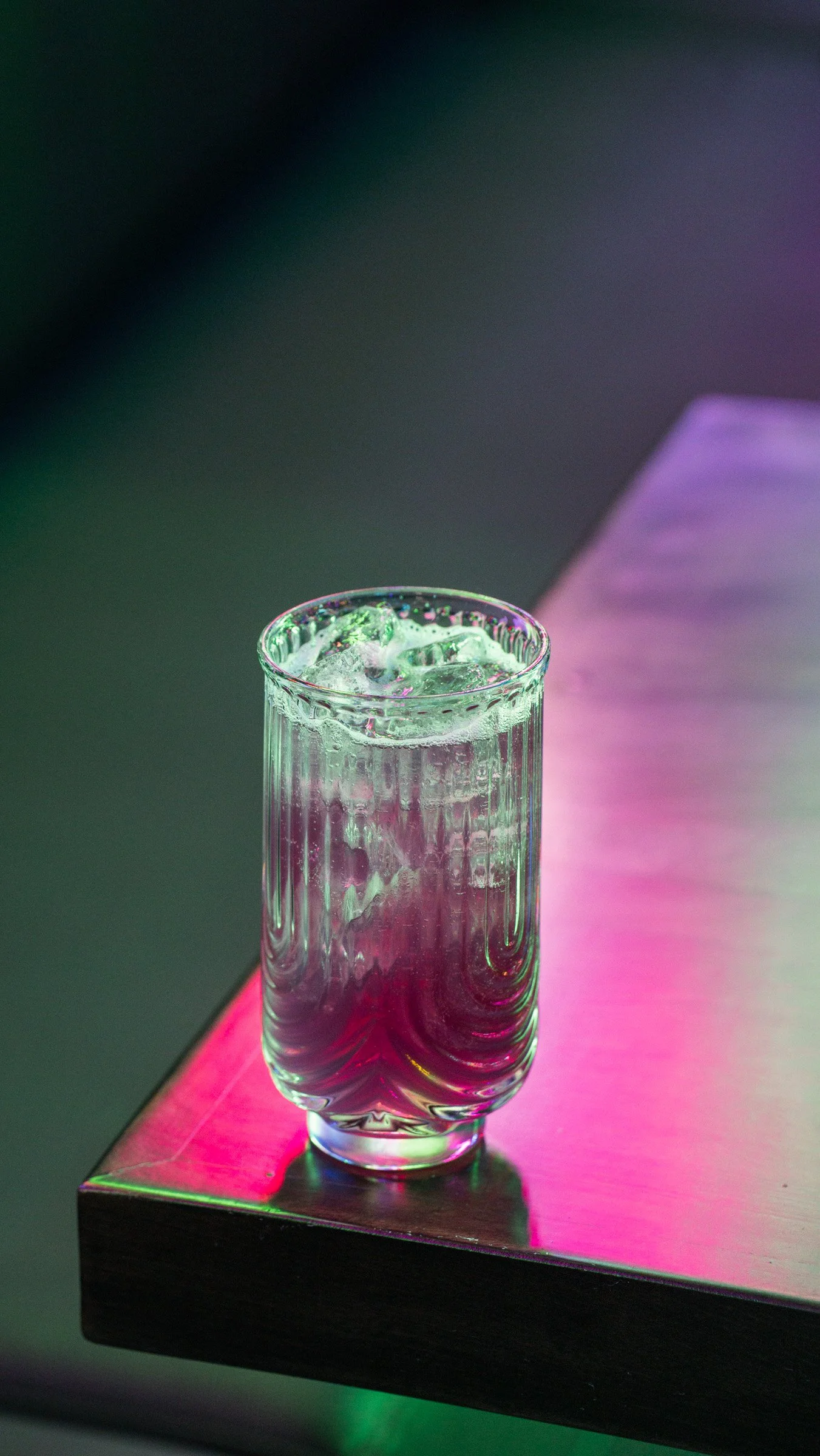 A clear glass filled with water and ice cubes, placed on a reflective surface with colorful lighting.