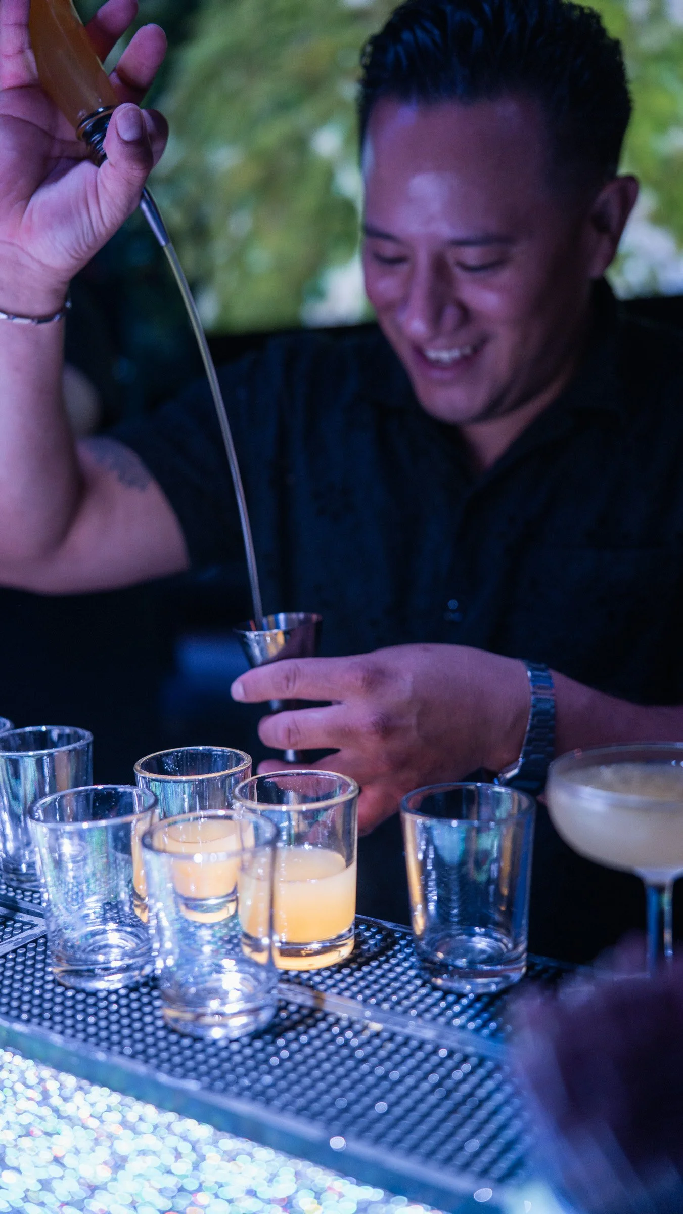 A man smiling as he uses a dropper to pour liquid into a shot glass surrounded by several other glasses on a bar counter.
