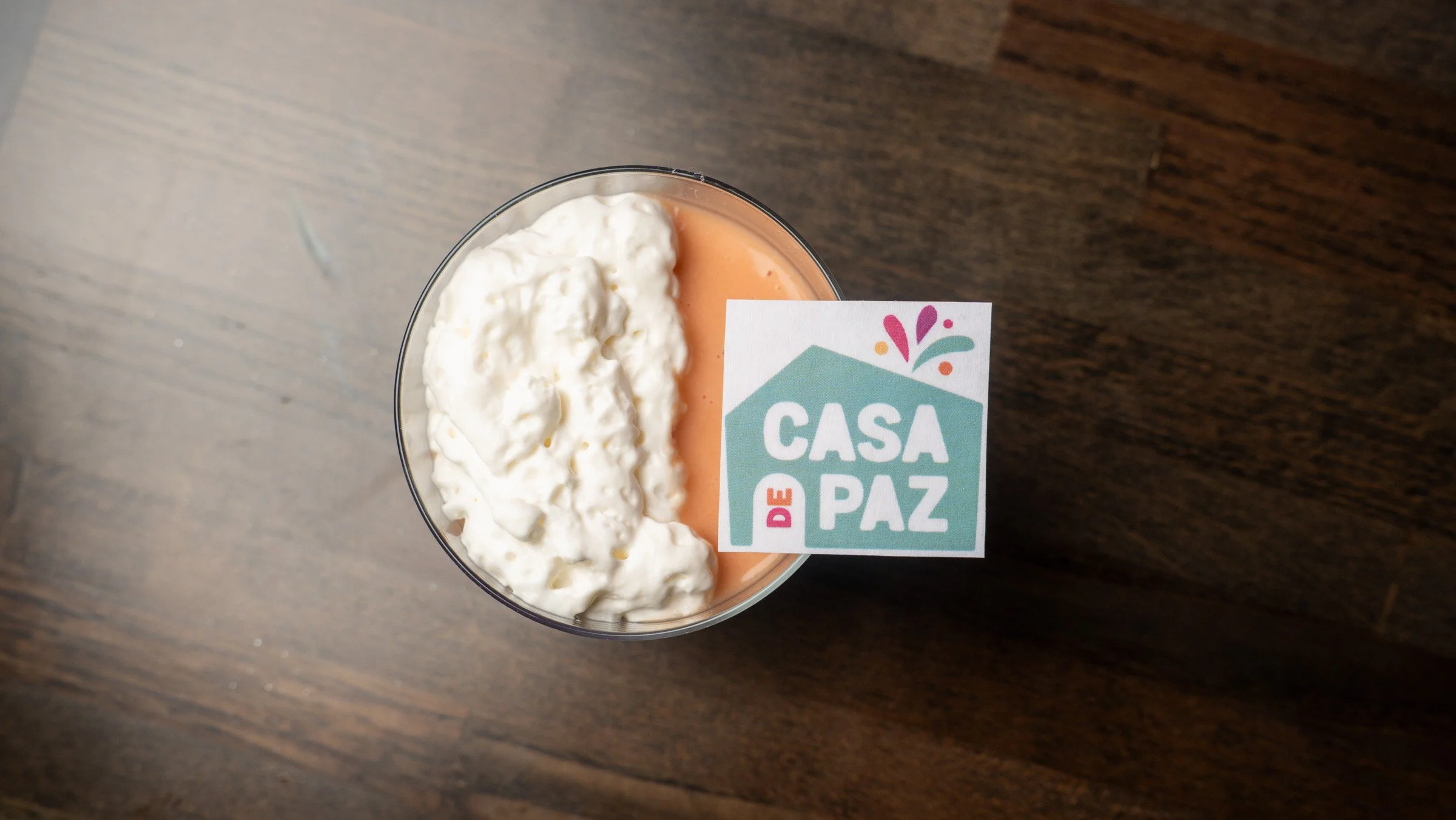 A clear plastic cup filled with two layers of creamy orange-colored beverage topped with whipped cream. A sticker with the words "Casa de Paz" and colorful design is attached to the cup. The cup is on a dark wooden surface.