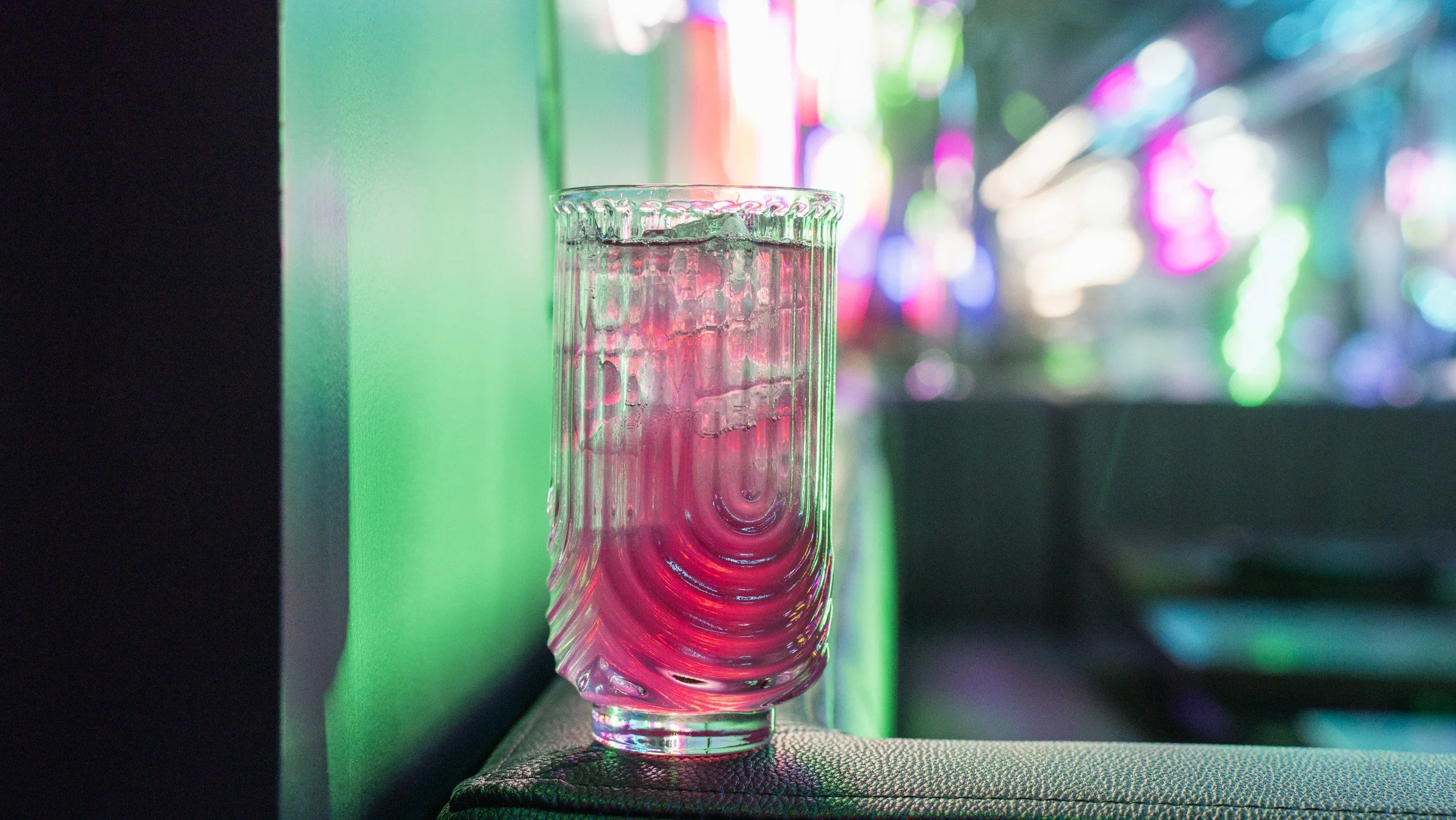 A tall, textured glass filled with pink liquid and ice, placed on a surface with vibrant, multicolored and blurry lighting in the background.