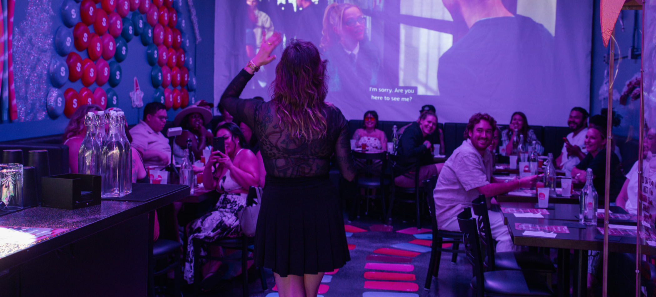 Inside a lively party venue with people sitting at tables, some smiling and taking photos. A woman with long curly hair and a black lace dress is standing and raising her hand. The venue has colorful decorations and a projector screen displaying a woman’s face and text.
