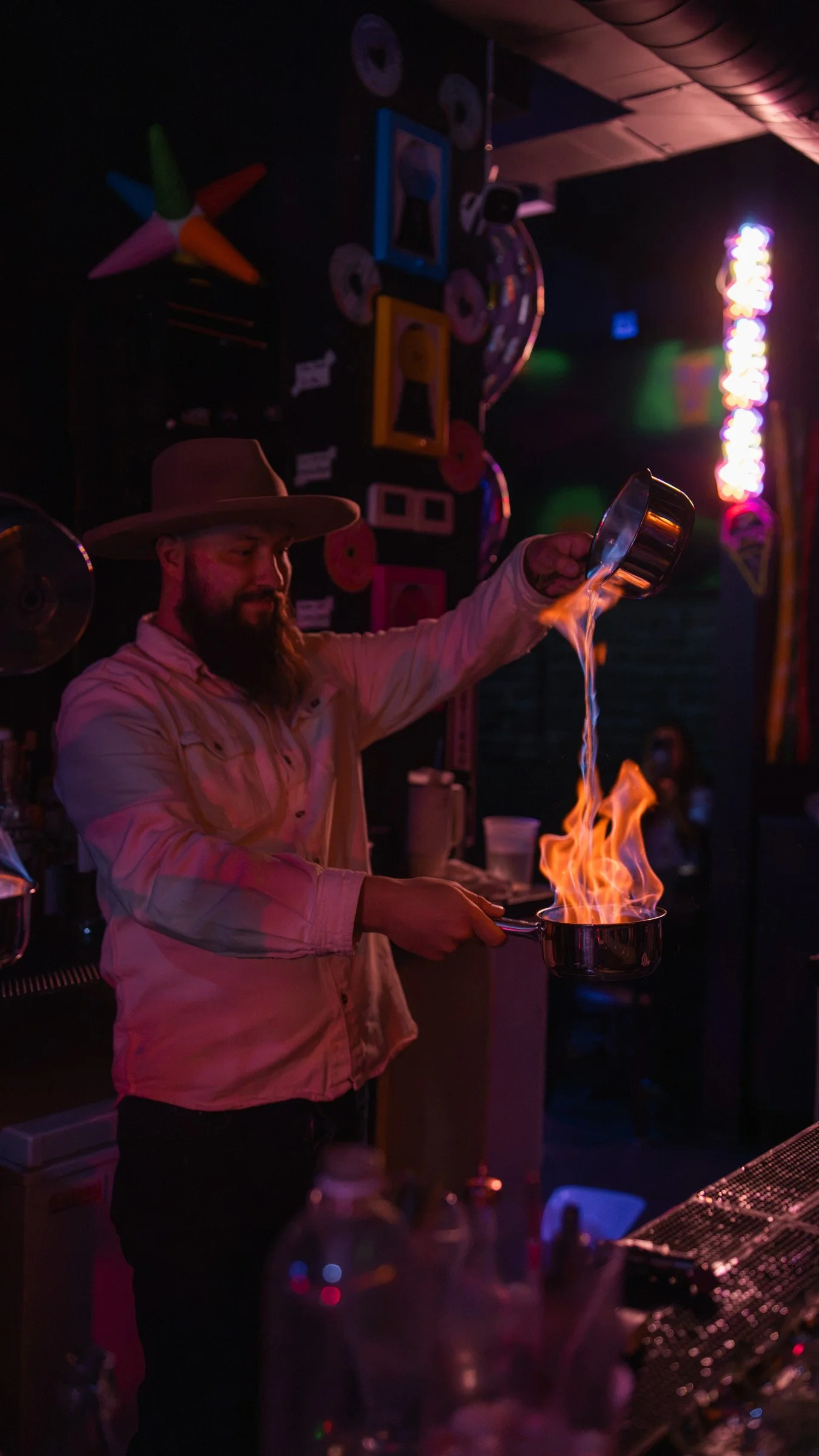 Bartender pouring liquid into a pan with flames, wearing a wide-brimmed hat, in a dimly lit bar with colorful decorations and neon lights.