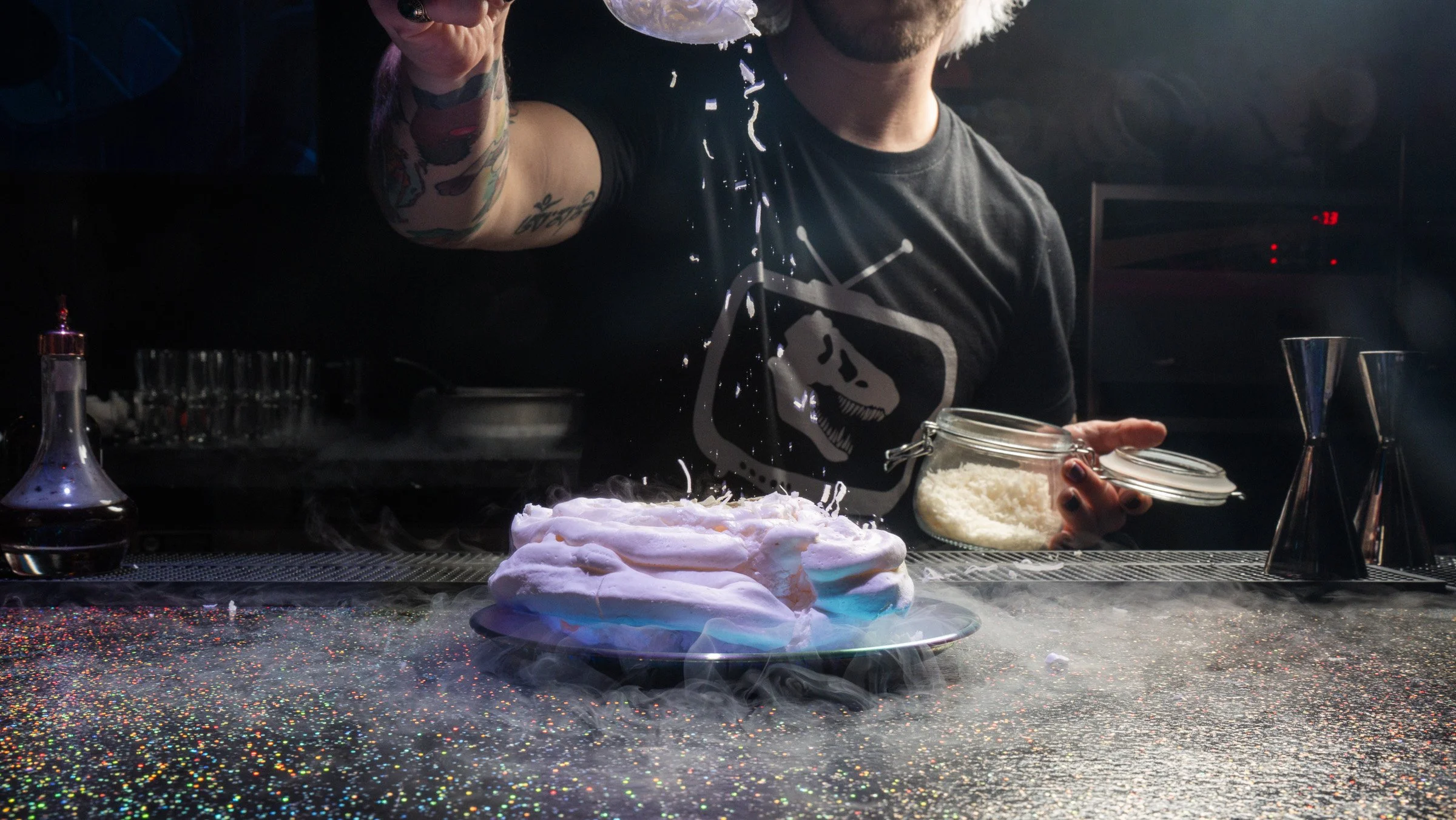 A man with tattoos, wearing a black shirt, is behind a counter with a spritzed cake in front of him. He is holding a jar of shredded white material, possibly cake toppings or icing. The background is dark with some equipment and a digital clock visible.