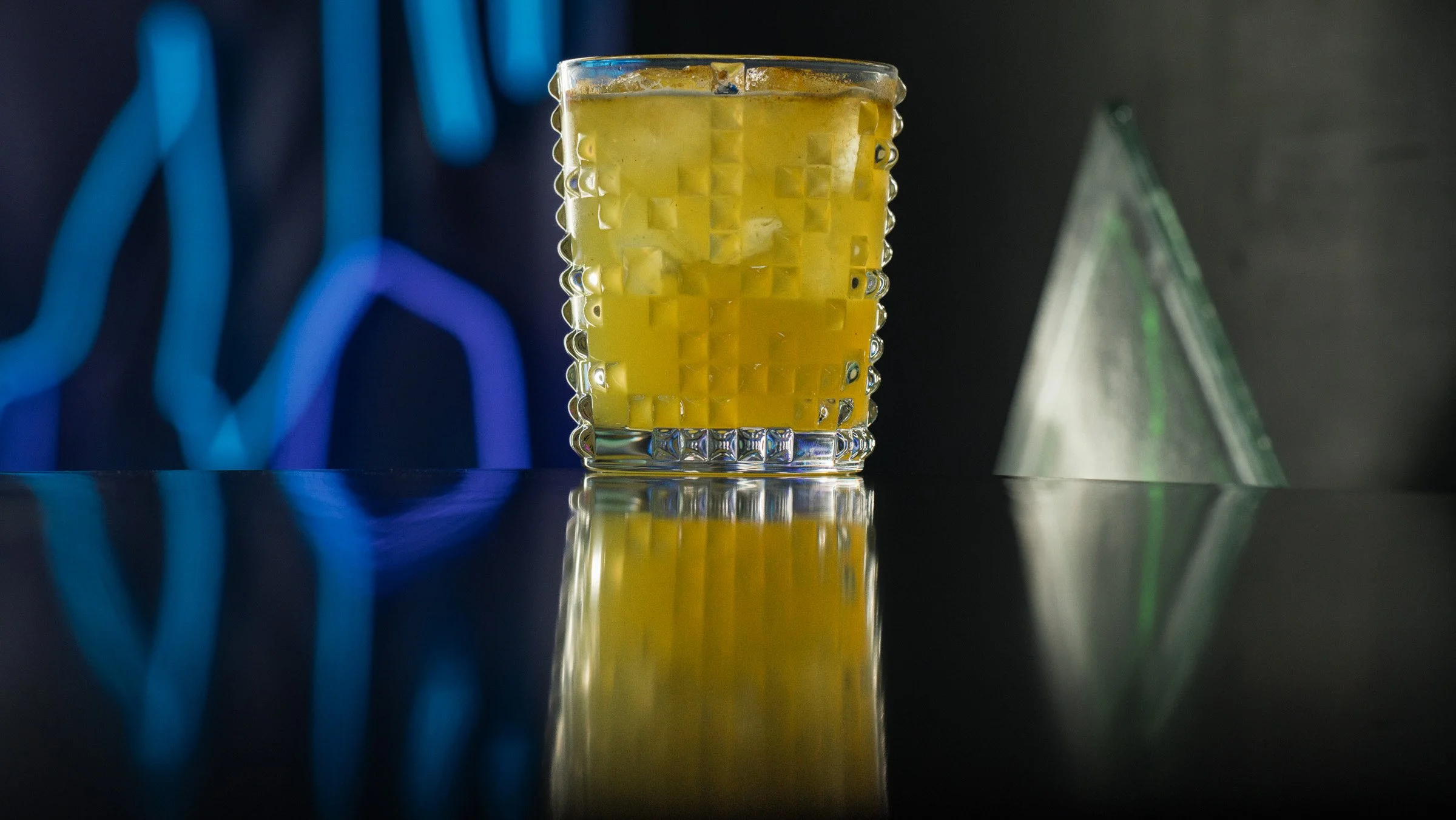 A glass of yellow liquid with ice cubes, placed on a reflective black surface, with abstract neon blue and green shapes in the background.