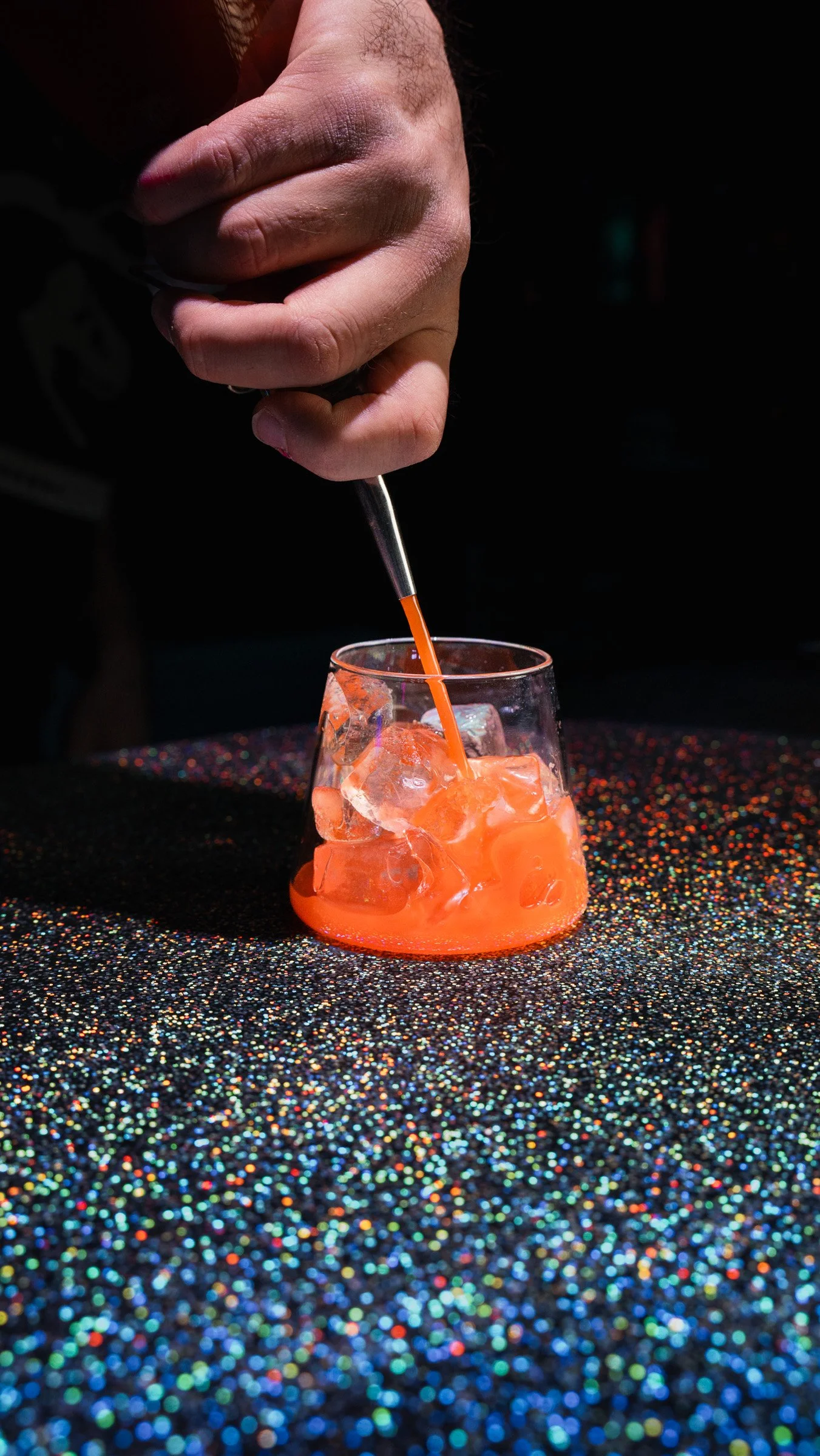 A hand holding a cocktail stirrer stirring an orange-colored drink with ice in a short glass on a glittery black surface.