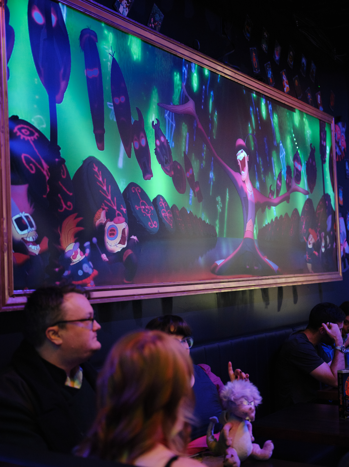 People sitting in a dark room watching a colorful animated display featuring a joyful skeleton and other characters with glowing symbols on rocks.