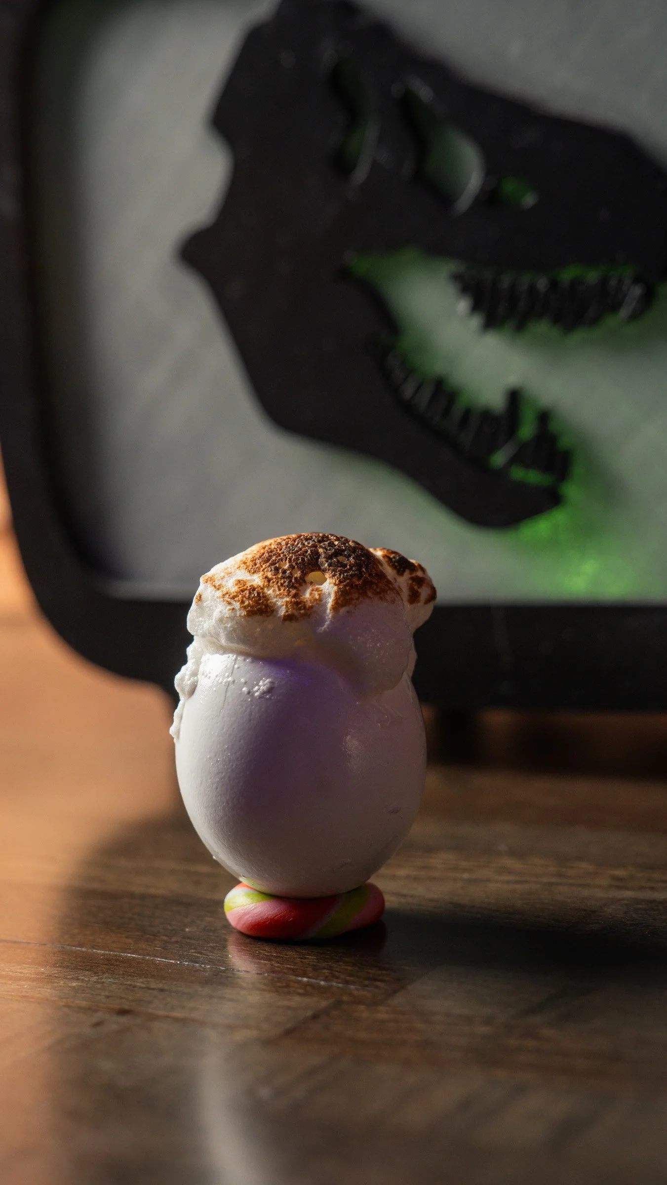 A scoop of vanilla ice cream topped with cocoa powder, standing on a small, multicolored base on a wooden surface. In the background, a dark tray with a chocolate dessert and green accents is partially visible.