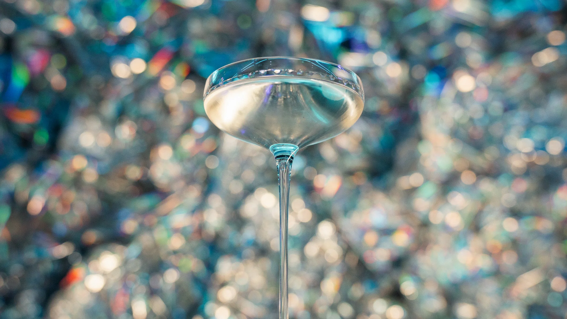 A cocktail glass with clear liquid on a table with a colorful, blurred background of shiny, reflective decorations.