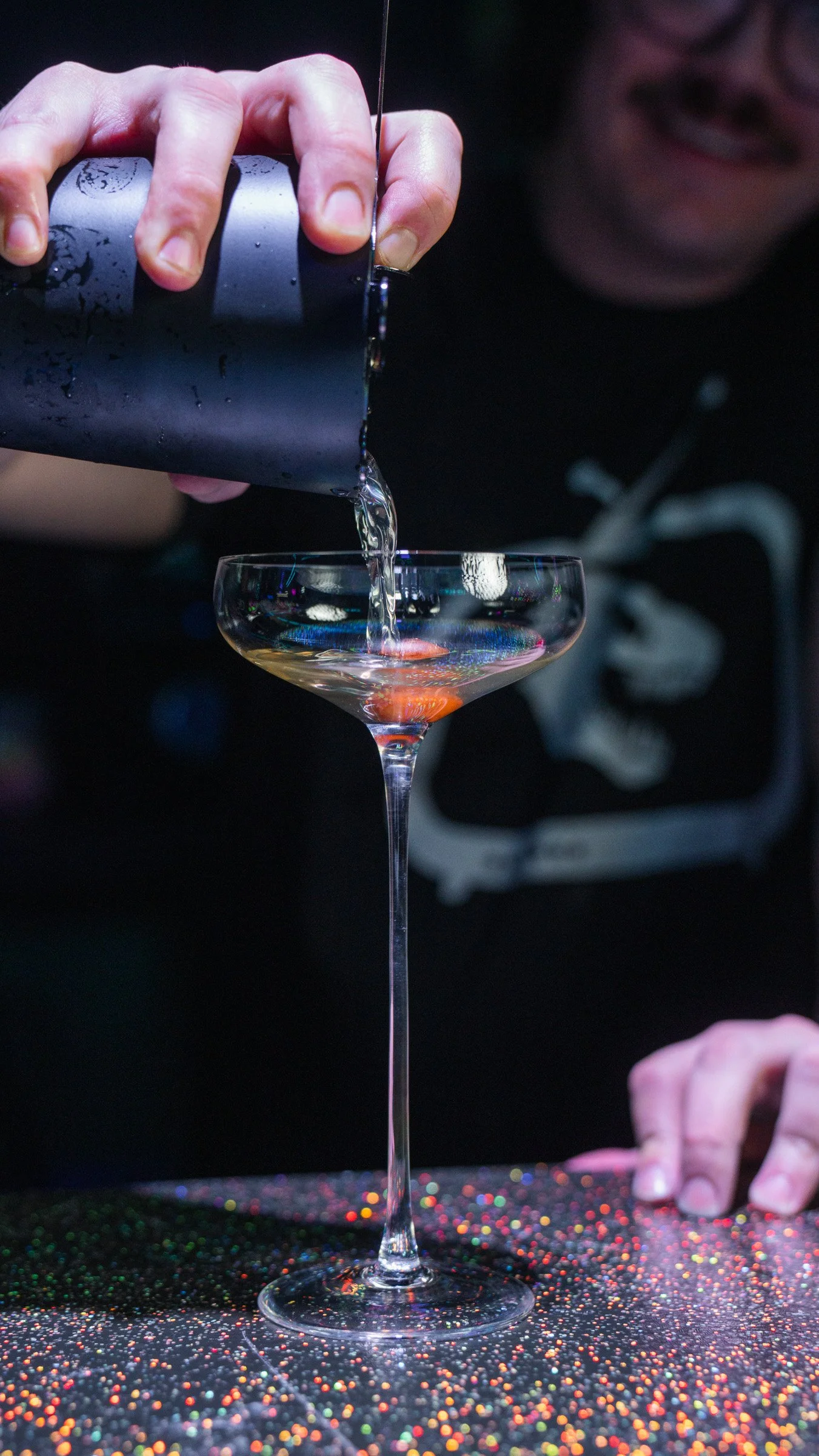 Person pouring a clear liquid into a cocktail glass, with a colorful, glittery surface underneath.