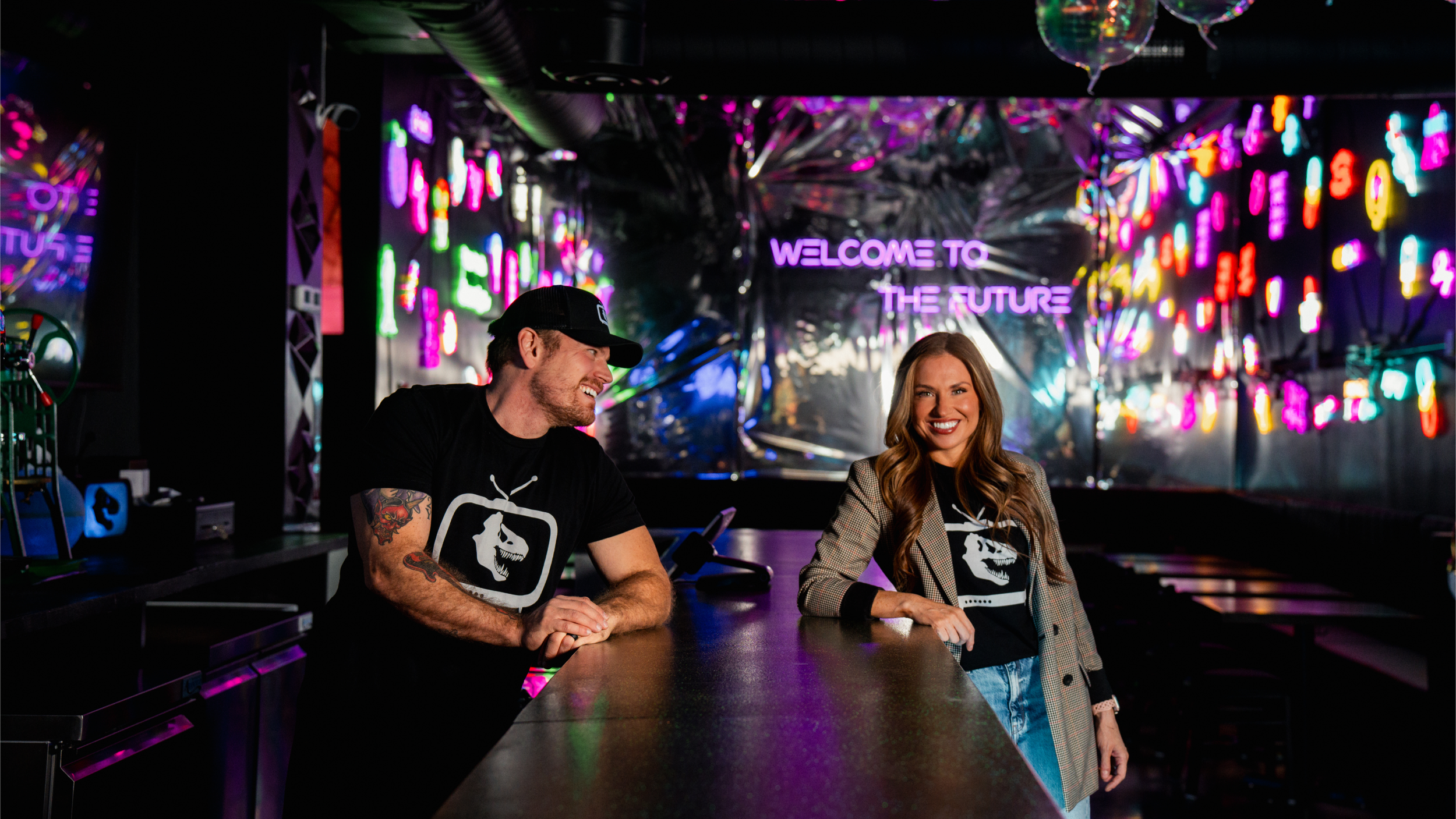 Two people, a man and a woman, sitting at a bar with a neon-lit futuristic background. The man is wearing a black cap and shirt with a logo, smiling and looking to his left. The woman is wearing a plaid blazer and black shirt with a logo, smiling and looking forward. The background features neon signs and the text "WELCOME TO THE FUTURE."