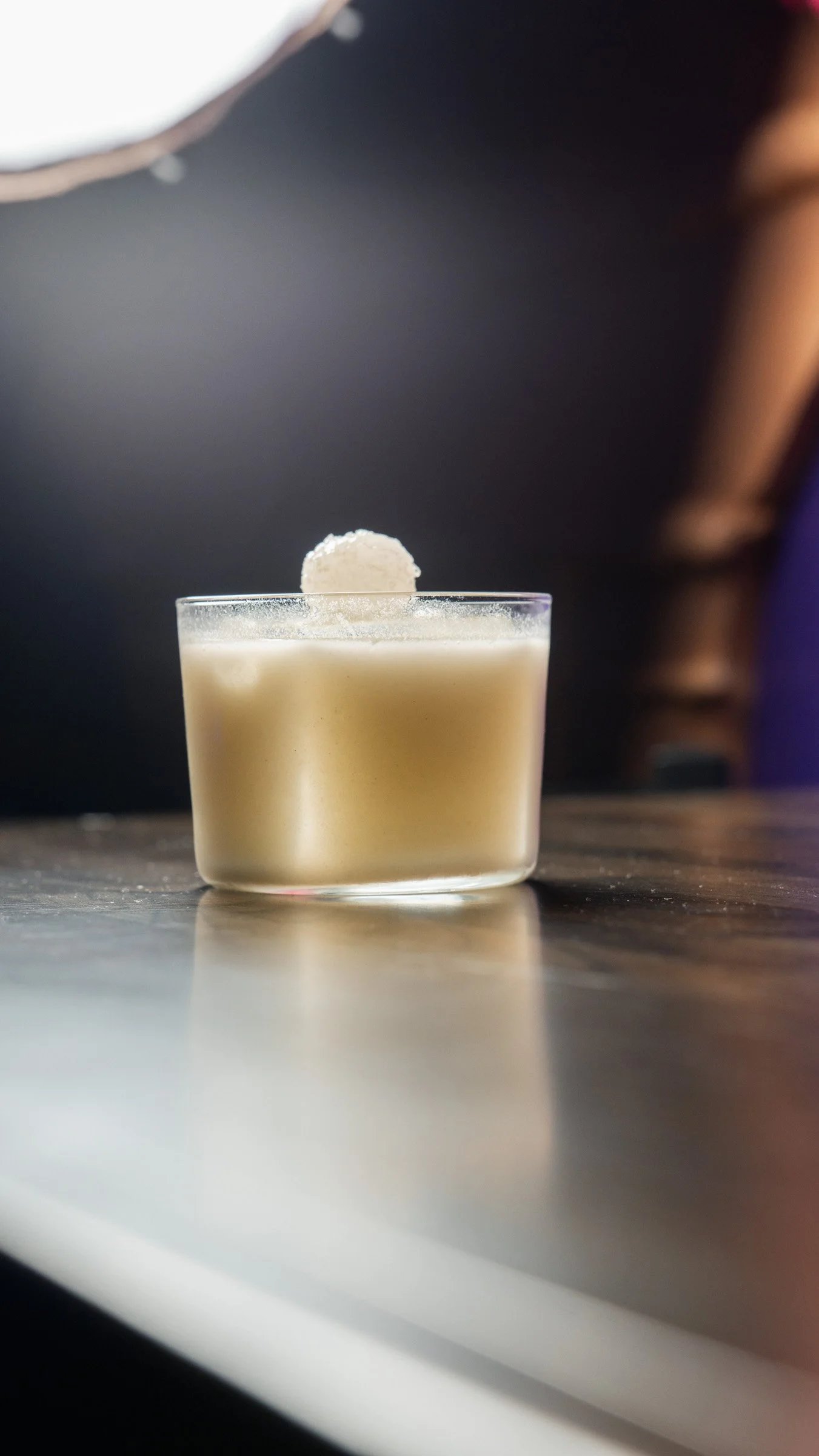 A glass container with a creamy, beige-colored substance and a small white ball-shaped garnish on top, placed on a reflective surface.