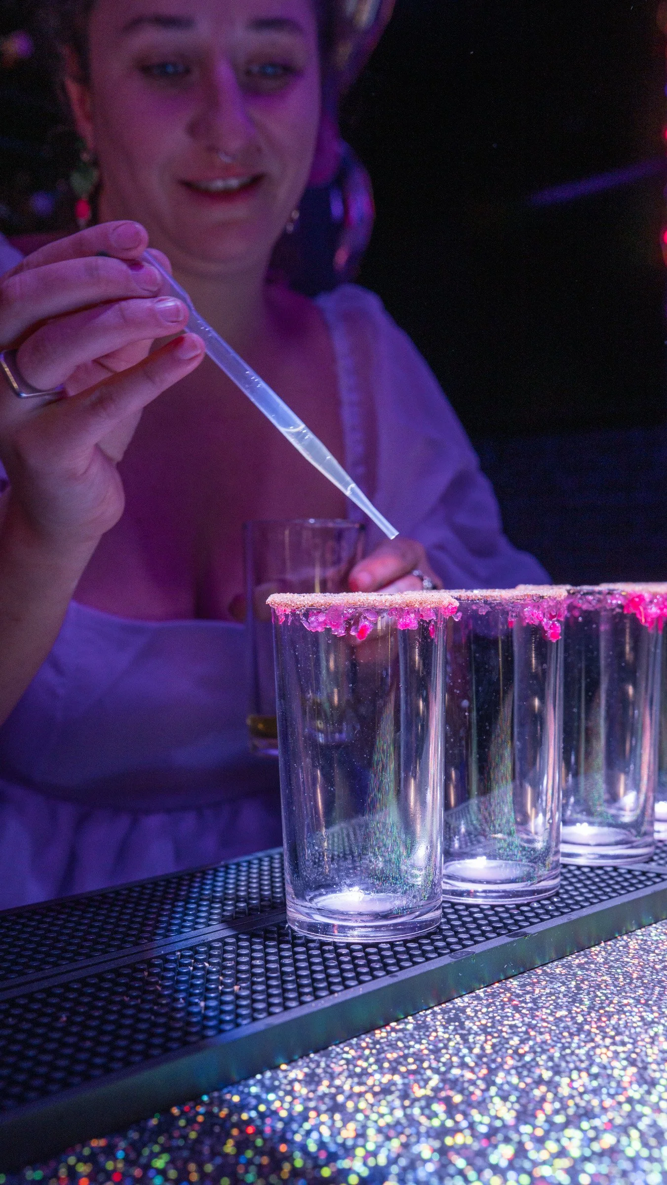 A woman is preparing drinks with a pipette in a bar or nightclub setting with colorful lighting. Several glasses with pink rim salt are on the bar counter, which is decorated with holographic glitter. The background is dark with hints of purple and red lights.