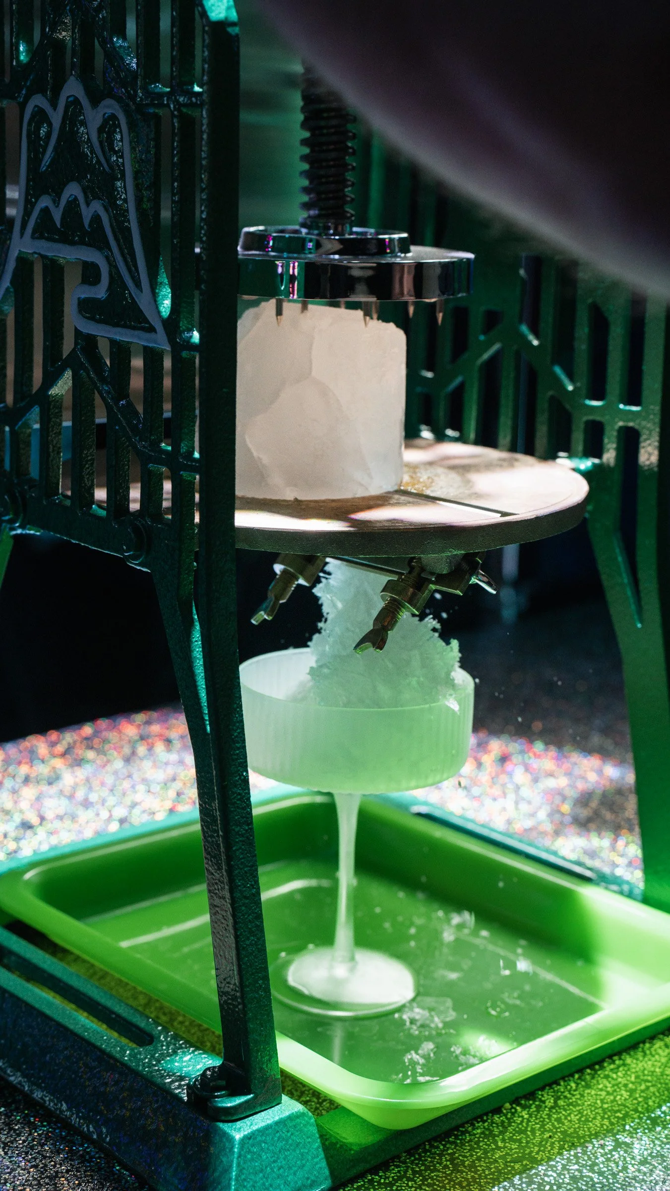 A science experiment setup with a metal frame, a block, and a mechanism dripping white liquid into a round container inside a green tray, with colorful glitter on the surface beneath.