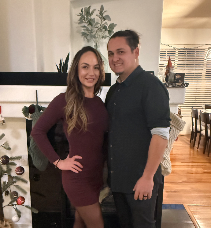 Two people smiling and posing together indoors, a woman in a maroon dress and a man in a black shirt, with a decorated fireplace and a Christmas tree in the background.