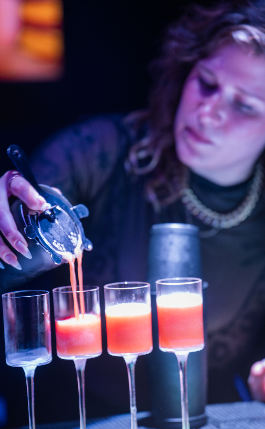 A bartender pouring a pink-colored cocktail into wine glasses at a bar.
