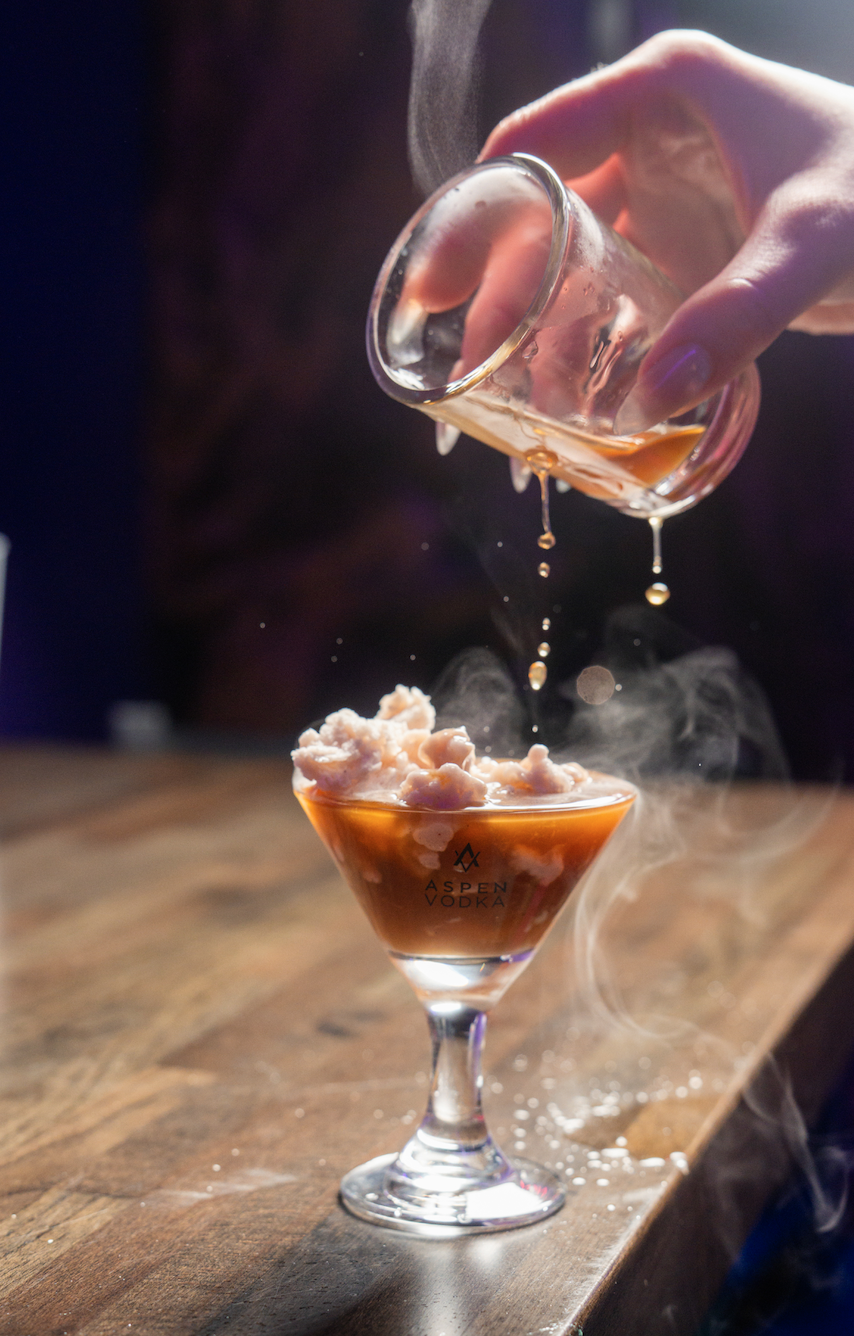 A hand pouring steaming liquid from a glass into a cocktail glass with ice cream inside, on a wooden table.