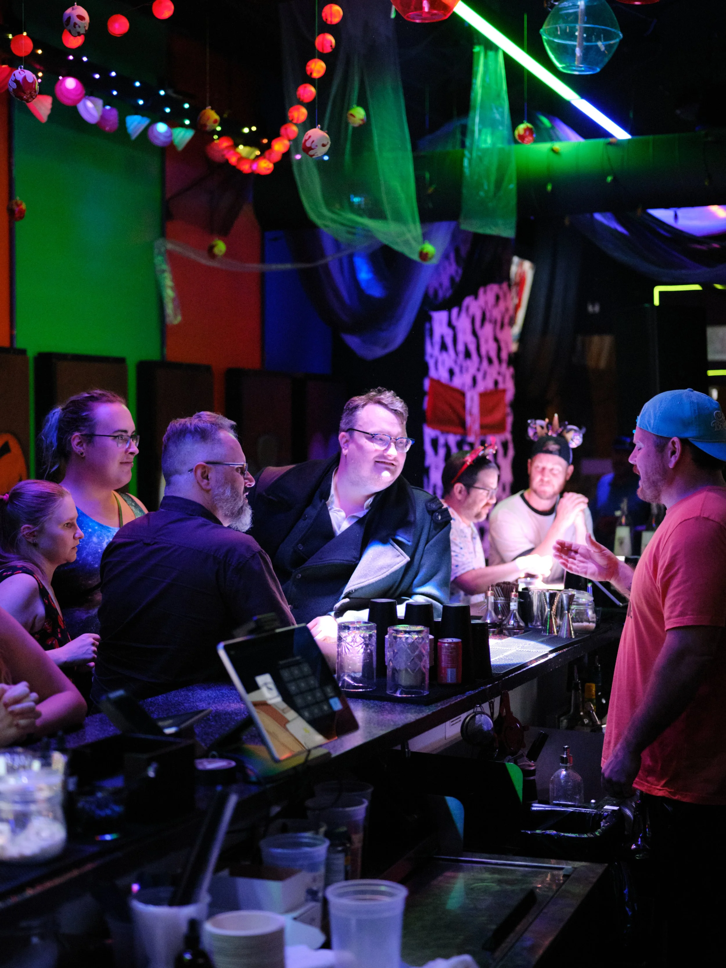 A group of people gathered at a bar with Halloween decorations hanging from the ceiling, including orange and purple skulls and red lantern-style lights. The bar area features various drinks, shakers, and bar equipment. The patrons are engaged in con