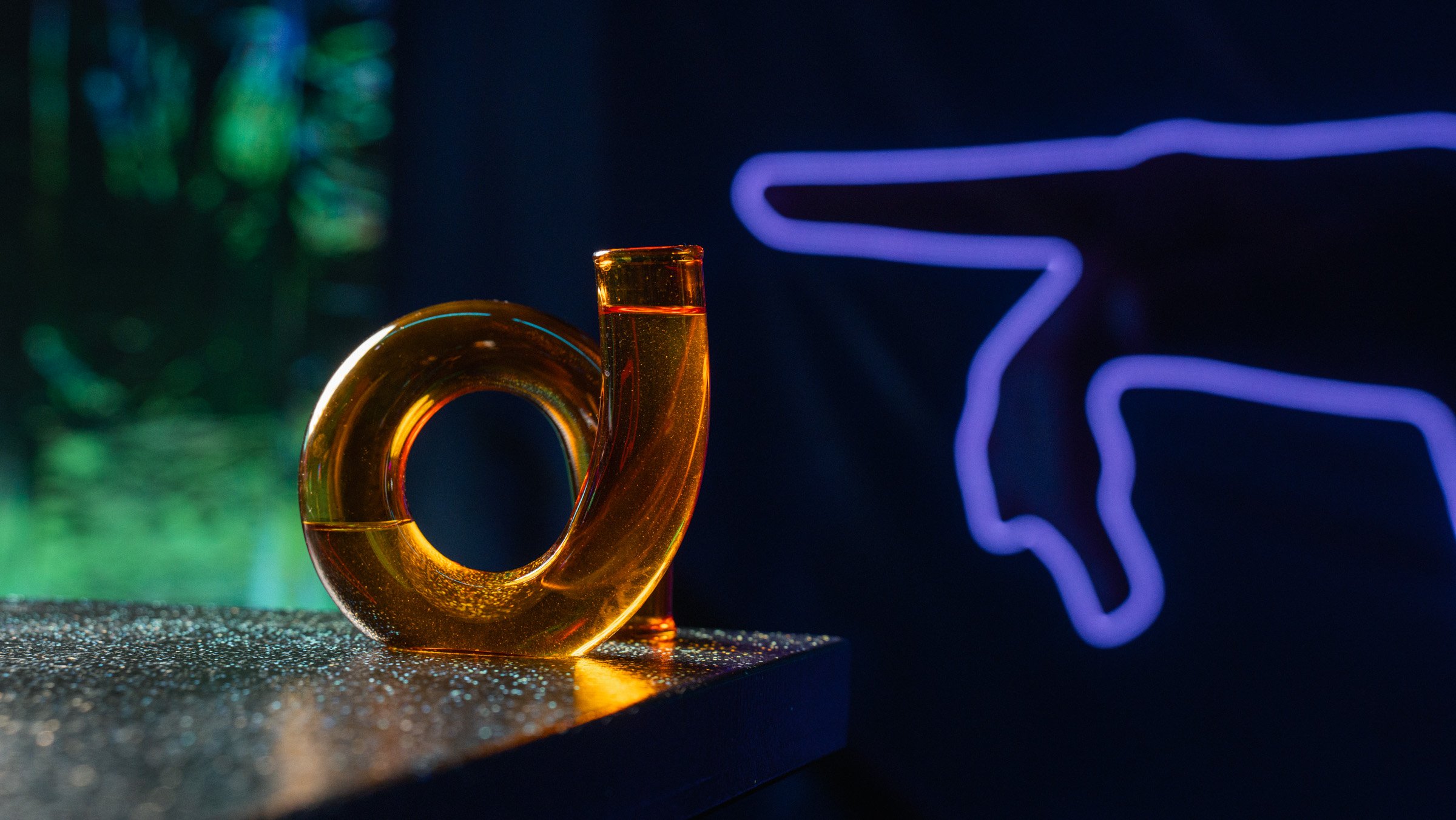A glass sculpture of a gold-colored number 0, illuminated by colorful lights, with a neon outline of an angel or cupid figure in blue in the background.