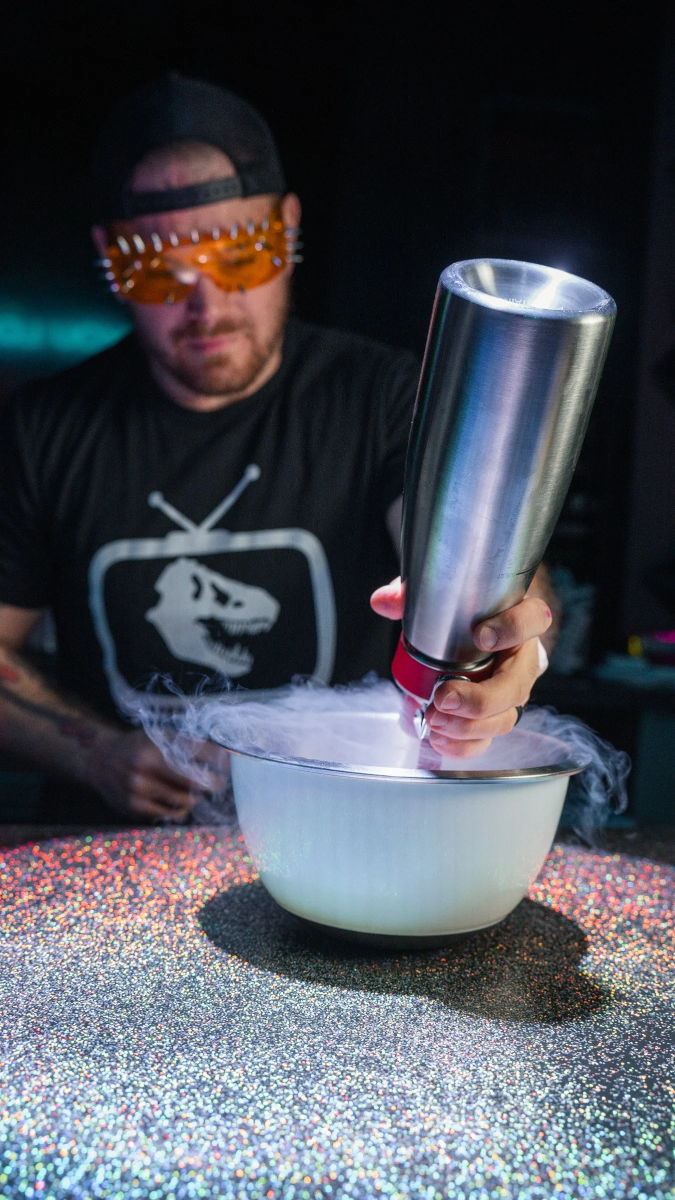 A man wearing orange science goggles and a black cap sprays something into a white bowl creating smoke or vapor, on a sparkly, multicolored surface.