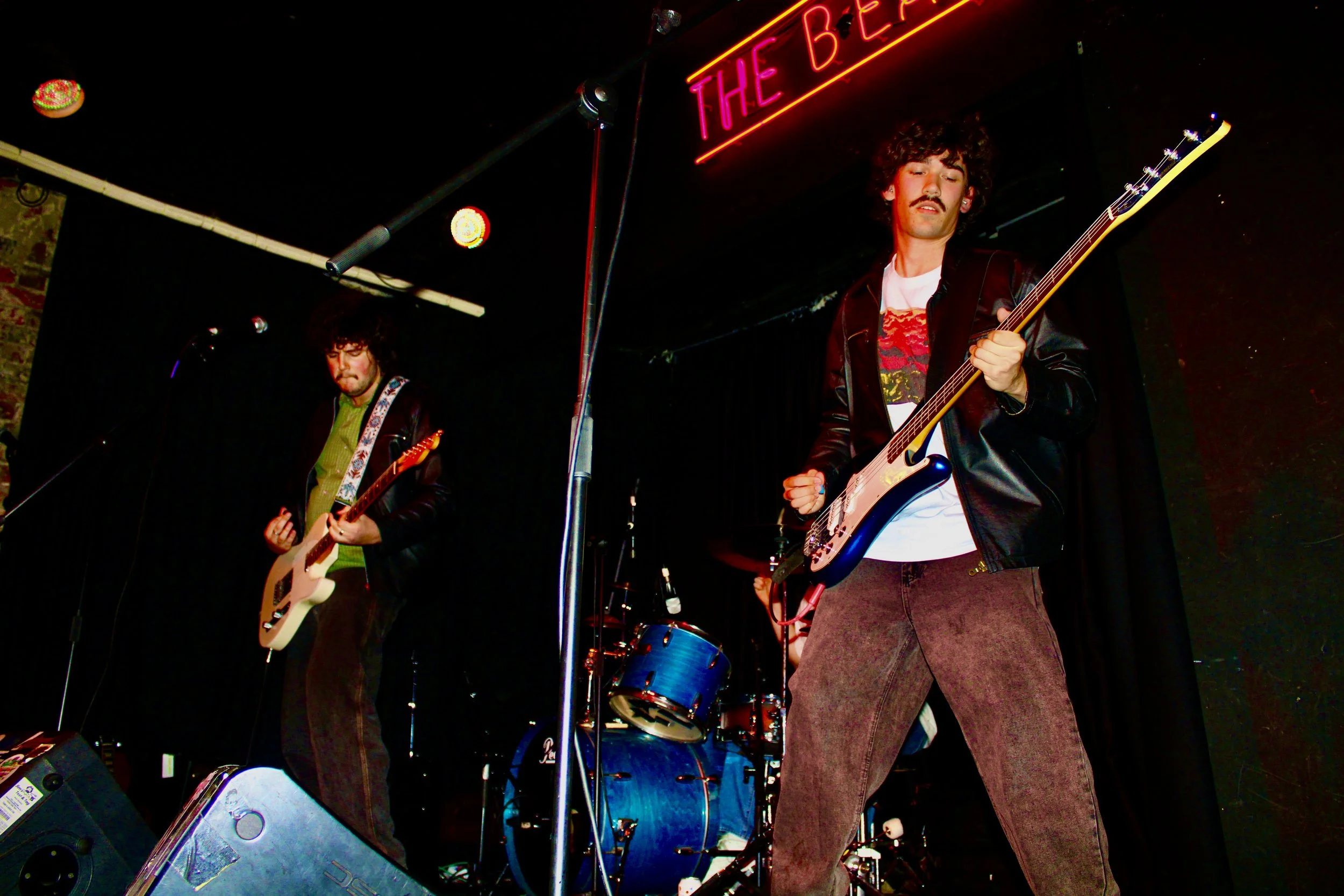 Two musicians playing electric guitars on stage with a drum set in the background. The stage has a dark backdrop and lit by colorful lights, with a red neon sign that reads 'THE BEER'.