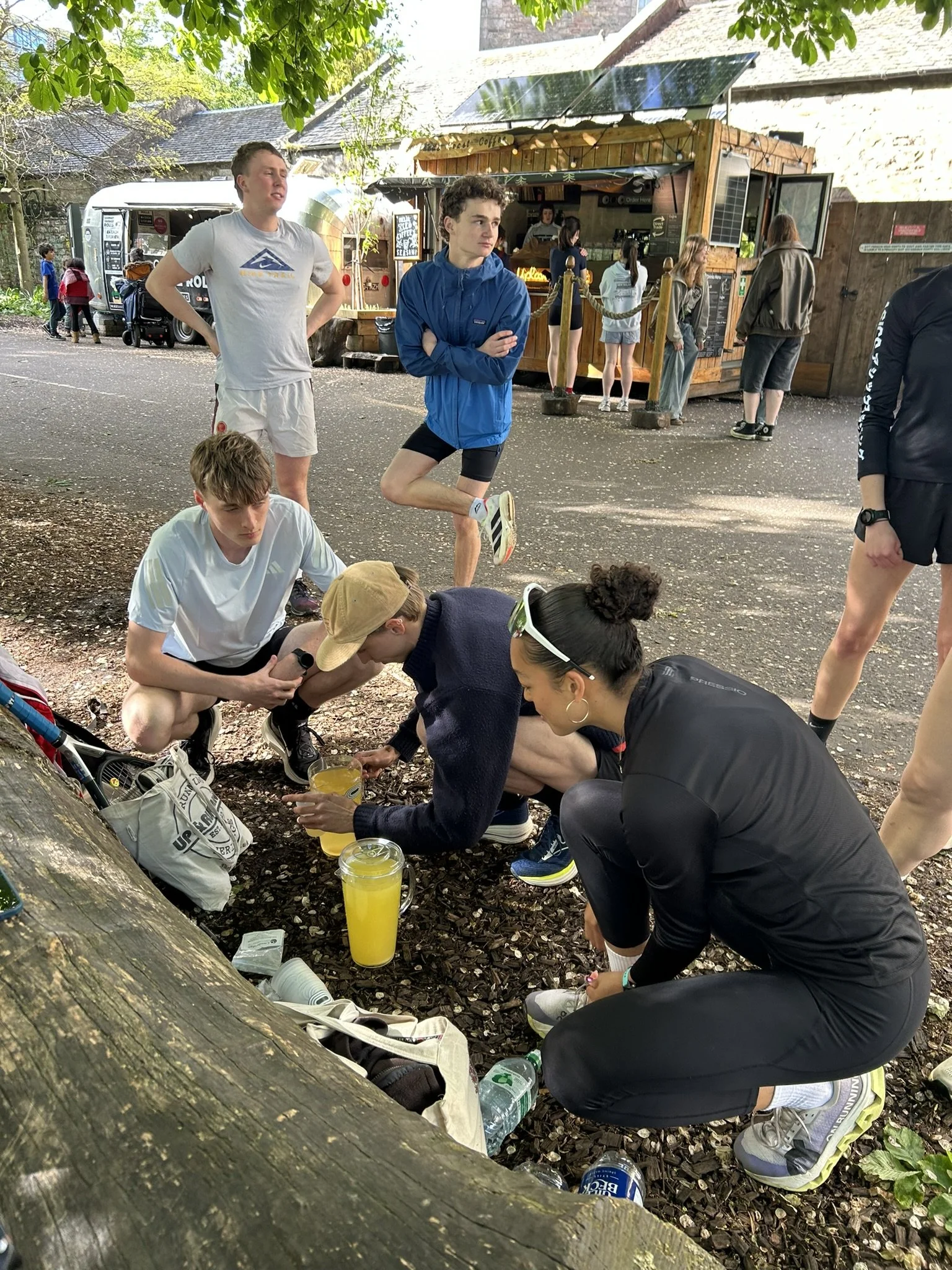 A group of young people gathering outdoors under a tree, some sitting and others standing, with a small booth or food stand in the background. Items like water bottles, cups with yellow drink, and personal belongings are visible on the ground.