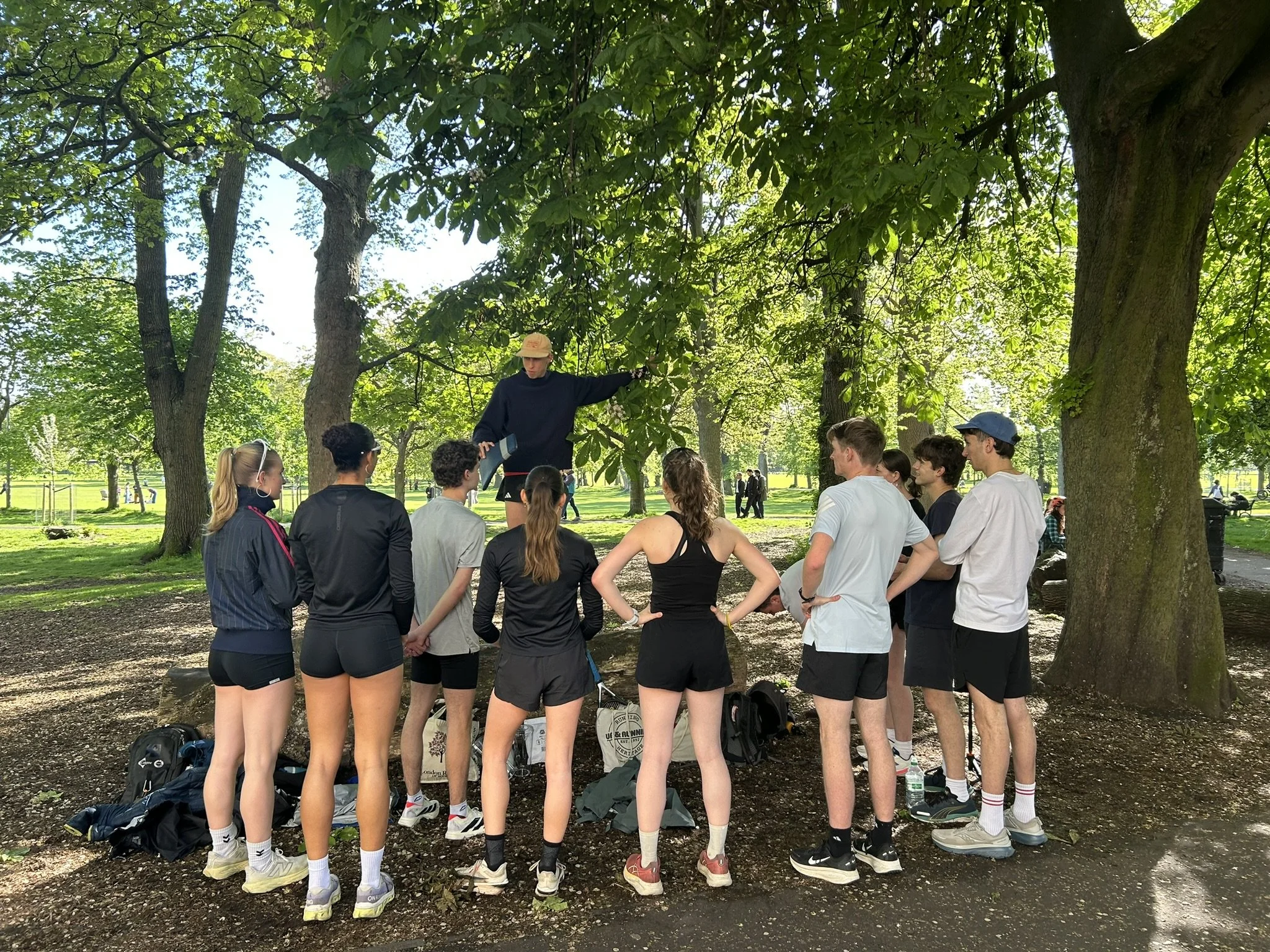 A group of people gathered outdoors in a park, listening to a person standing on a raised surface, possibly giving instructions or speaking. The group includes mostly young women and men dressed in athletic clothing, with trees and green space in the