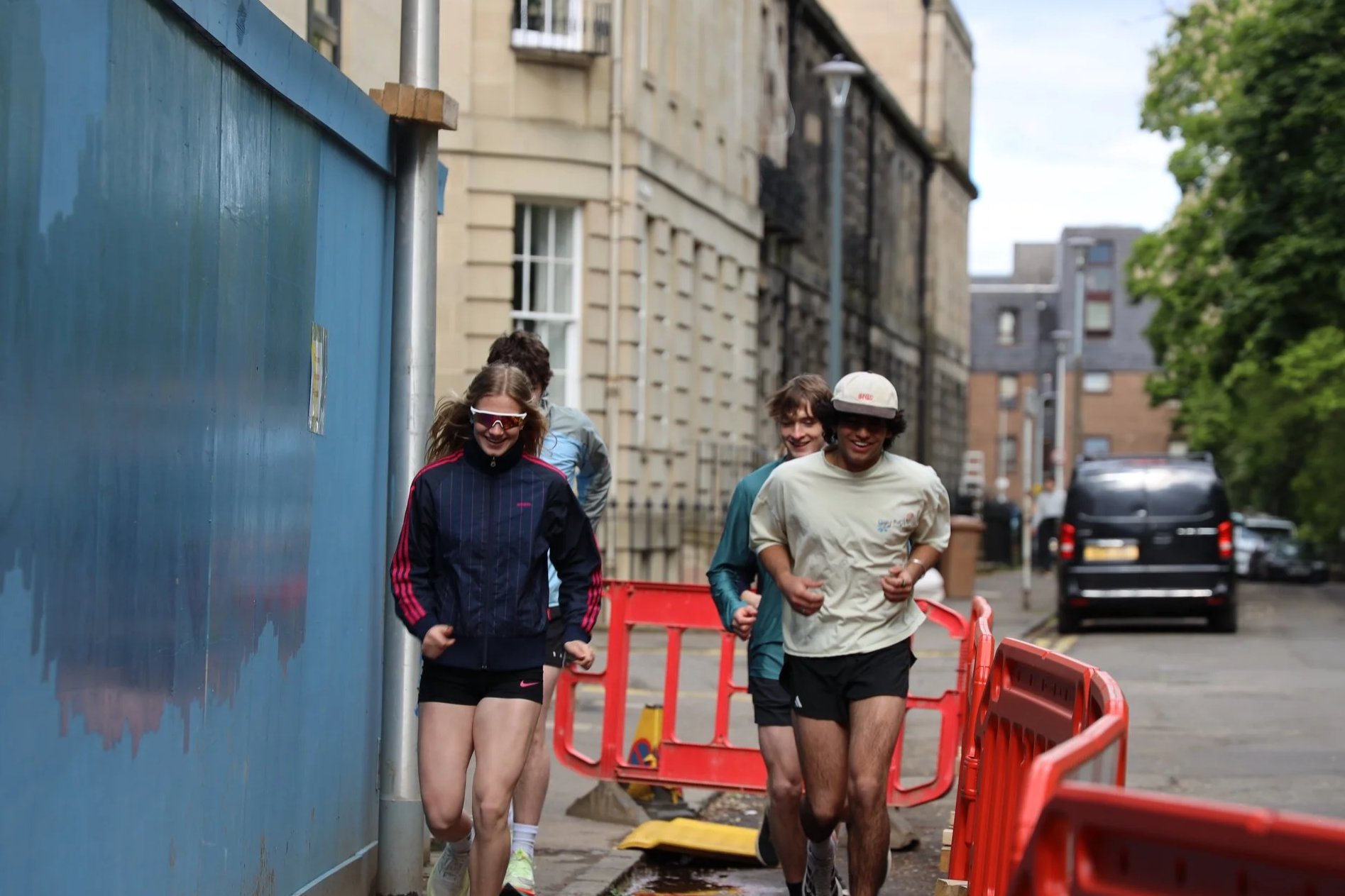 A group of four young people jogging on a city street behind orange barriers, with a blue wall on the left and buildings on the right, during daytime.