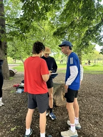 Three young men standing outdoors under a tree, engaged in a conversation, with a park background and some other people visible in the distance.
