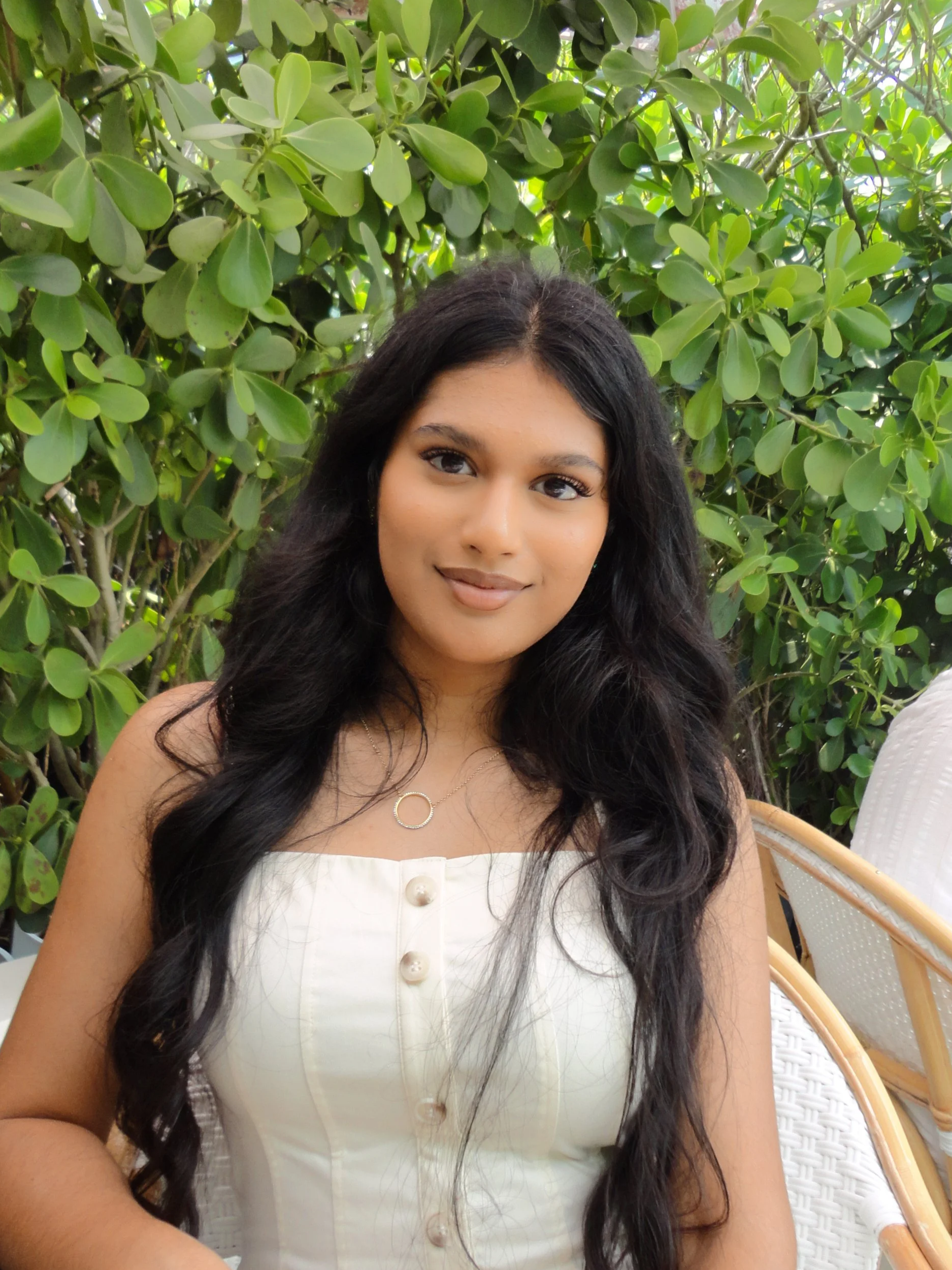 A woman with long black wavy hair and medium skin tone sitting outdoors in front of green leafy bushes, wearing a white sleeveless top with pearl buttons and a necklace with a circular pendant.