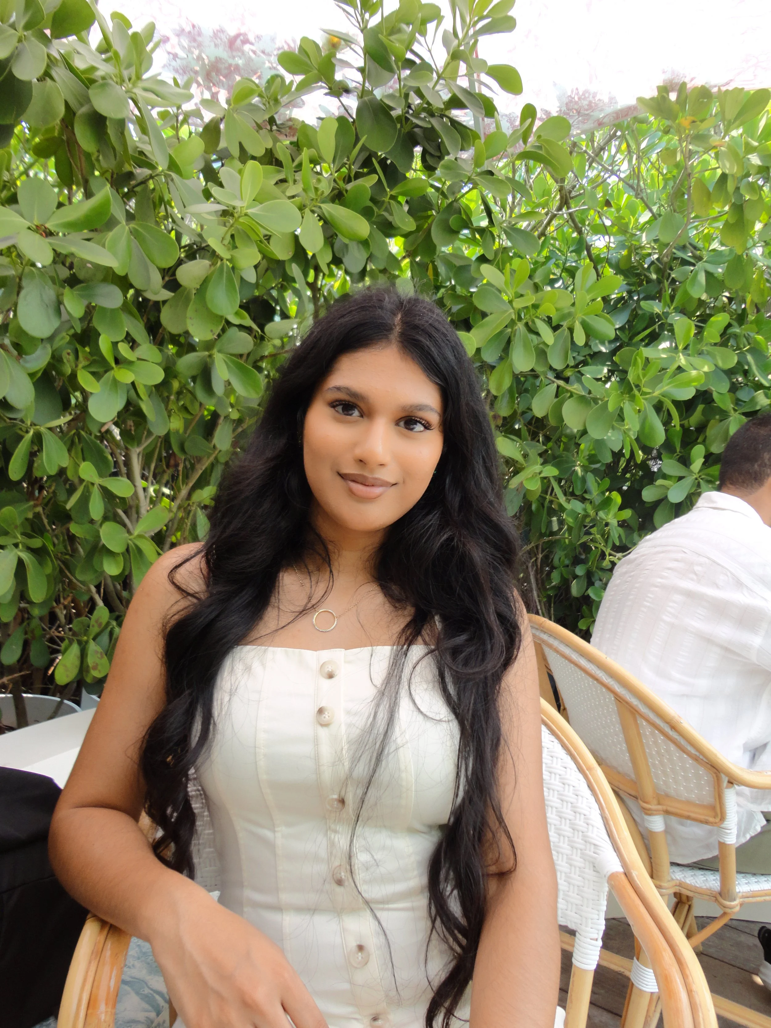 A young woman with long black hair and light brown skin sitting at a table outdoors, wearing a white sleeveless top with buttons and a circular necklace, surrounded by green leafy plants.