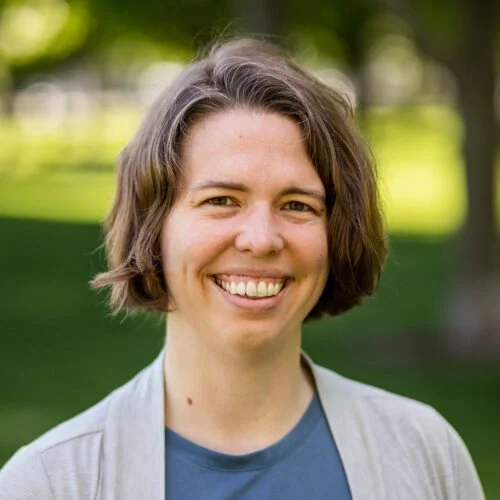 Woman posing with short brown hair, blue shirt with a light grey cardigan, and smiling