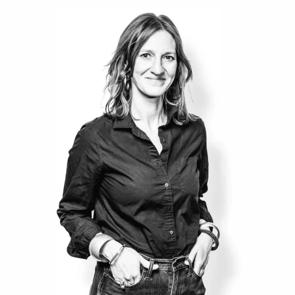 Black and white photo of woman with shoulder-length hair, collared dark shirt, smiling and posing at camera with hands in jean pockets