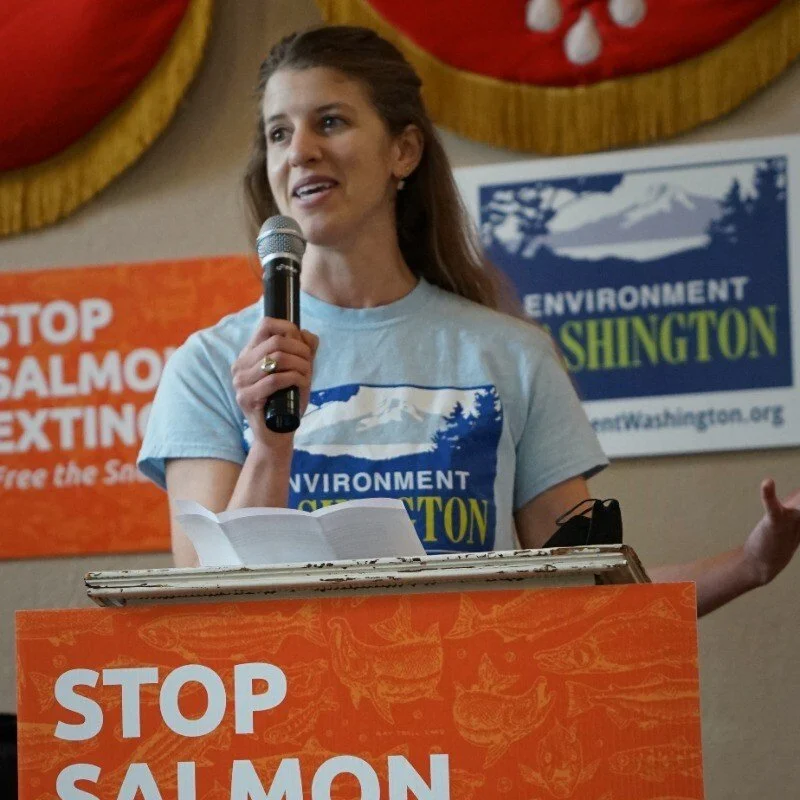 Woman with a light blue shirt, long brown hair, standing in front of a podium with a mic in her hand speaking