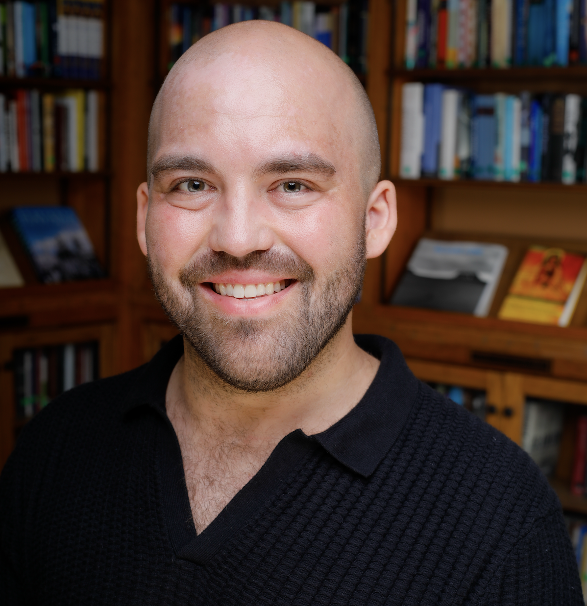 Male with beard and bald head, posing and smiling in a collared v-neck black shirt