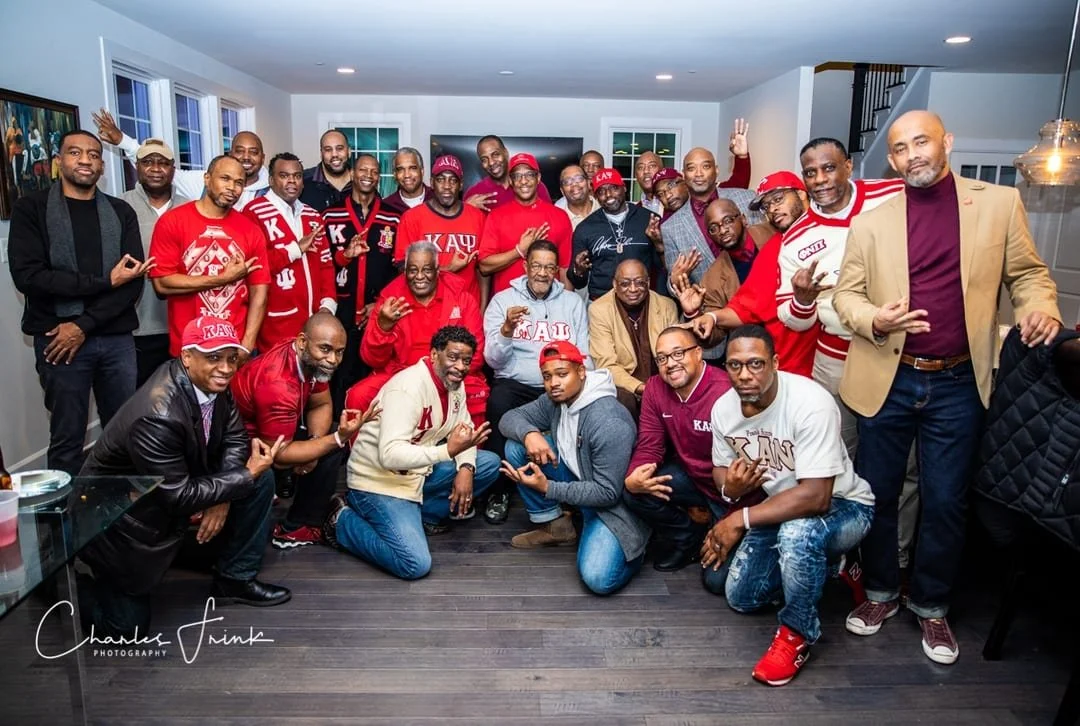Group of men gathered indoors, some wearing red clothing and hats with Greek letters, posing for a photo and making hand gestures.